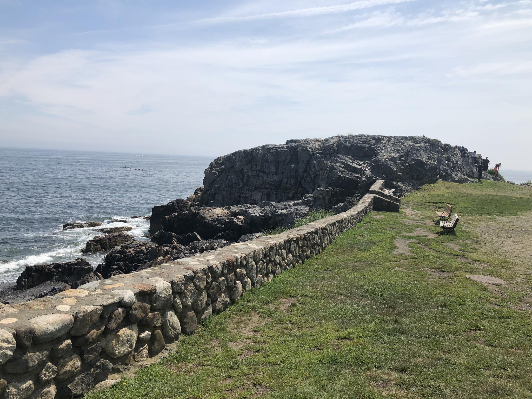 Rocky coastline with a stone wall, grassy cliff, and ocean on a sunny day.