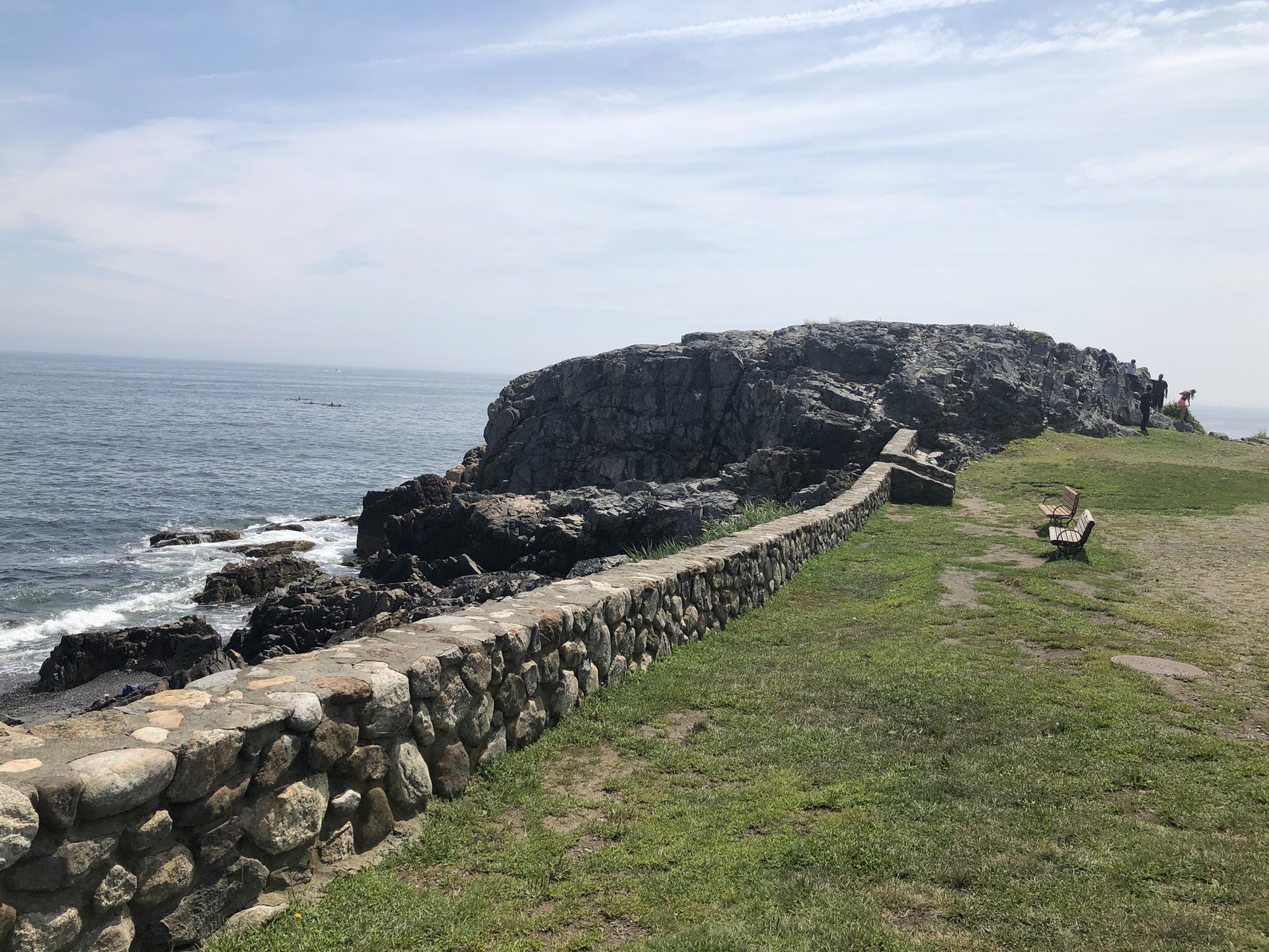 Rocky coastline with a stone wall, grassy cliff, and ocean on a sunny day.