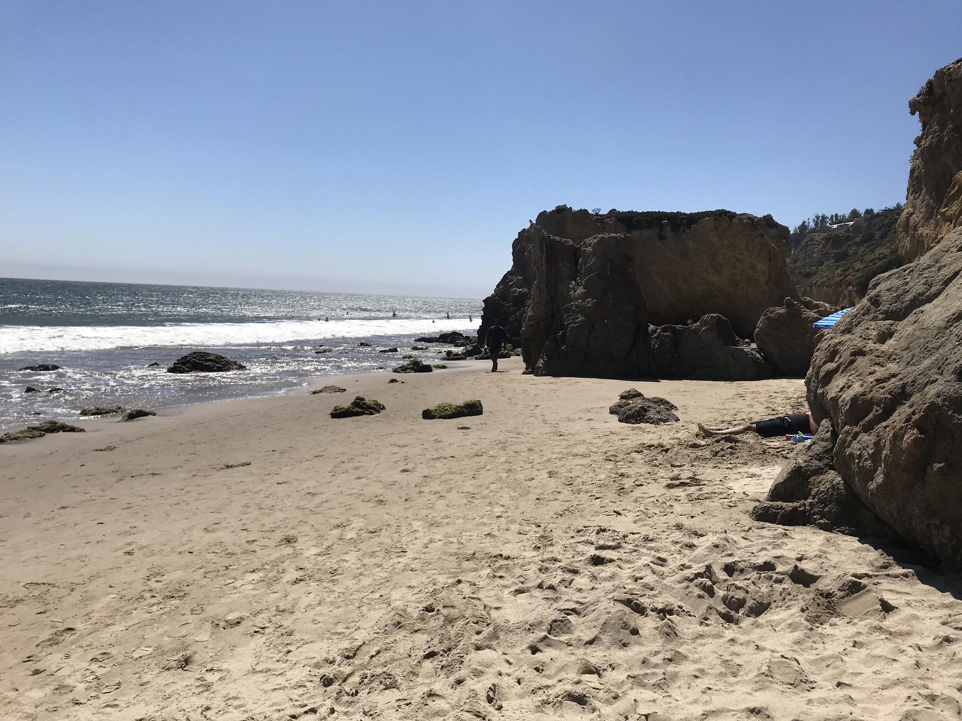 Sandy beach with large rock formations, ocean waves, and distant people under bright sun.