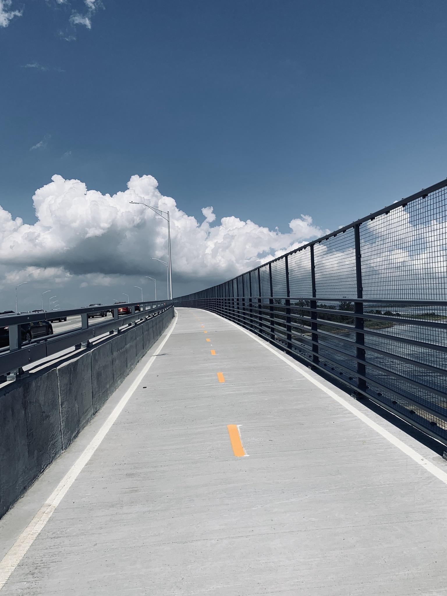 Paved pedestrian bridge with metal fence under bright blue sky with clouds.