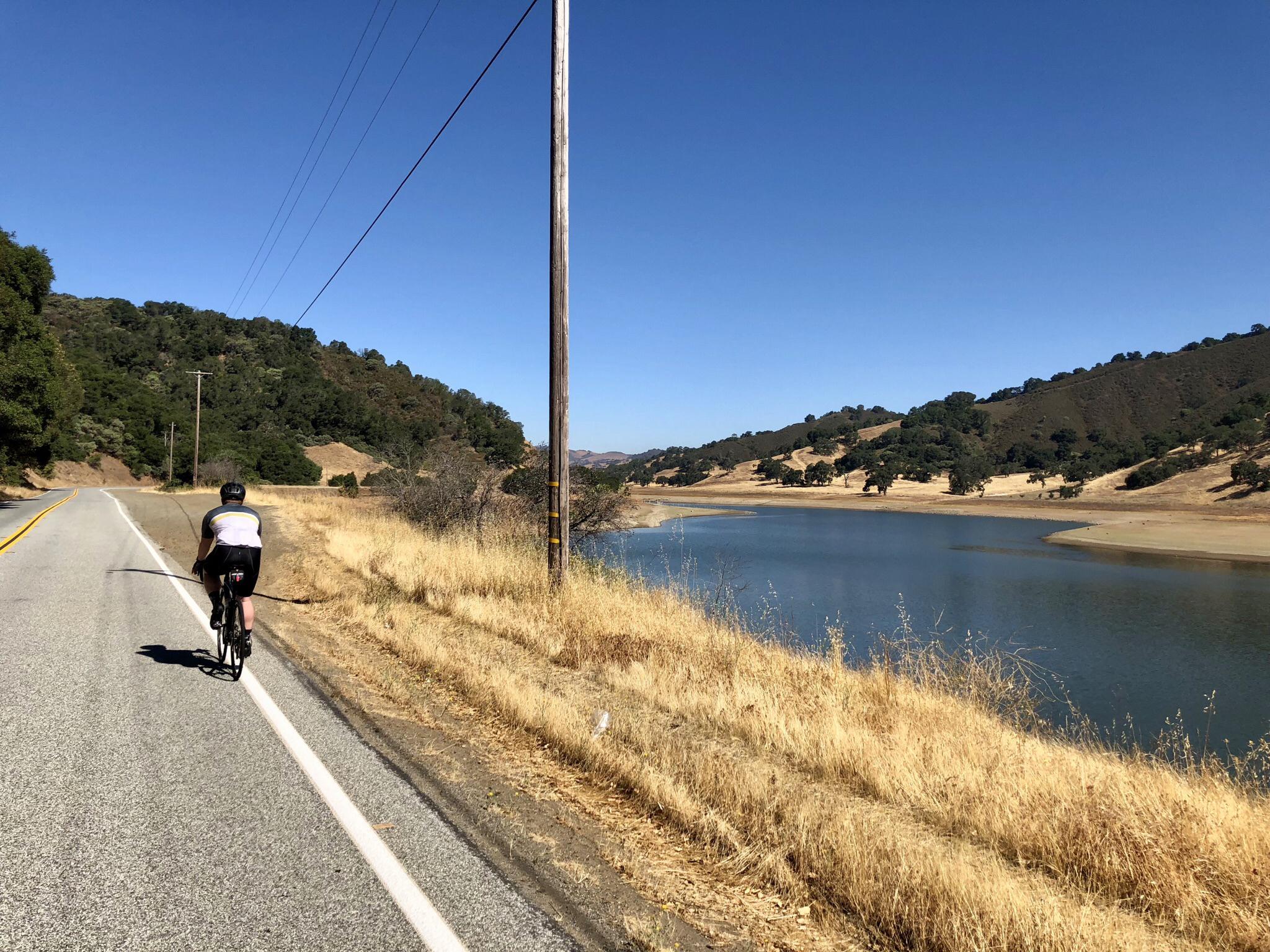 A cyclist rides along a road next to a lake under a clear sky.