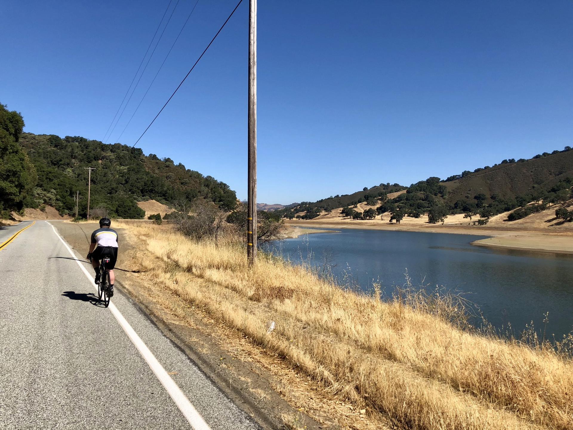 A cyclist rides along a road next to a lake under a clear sky.