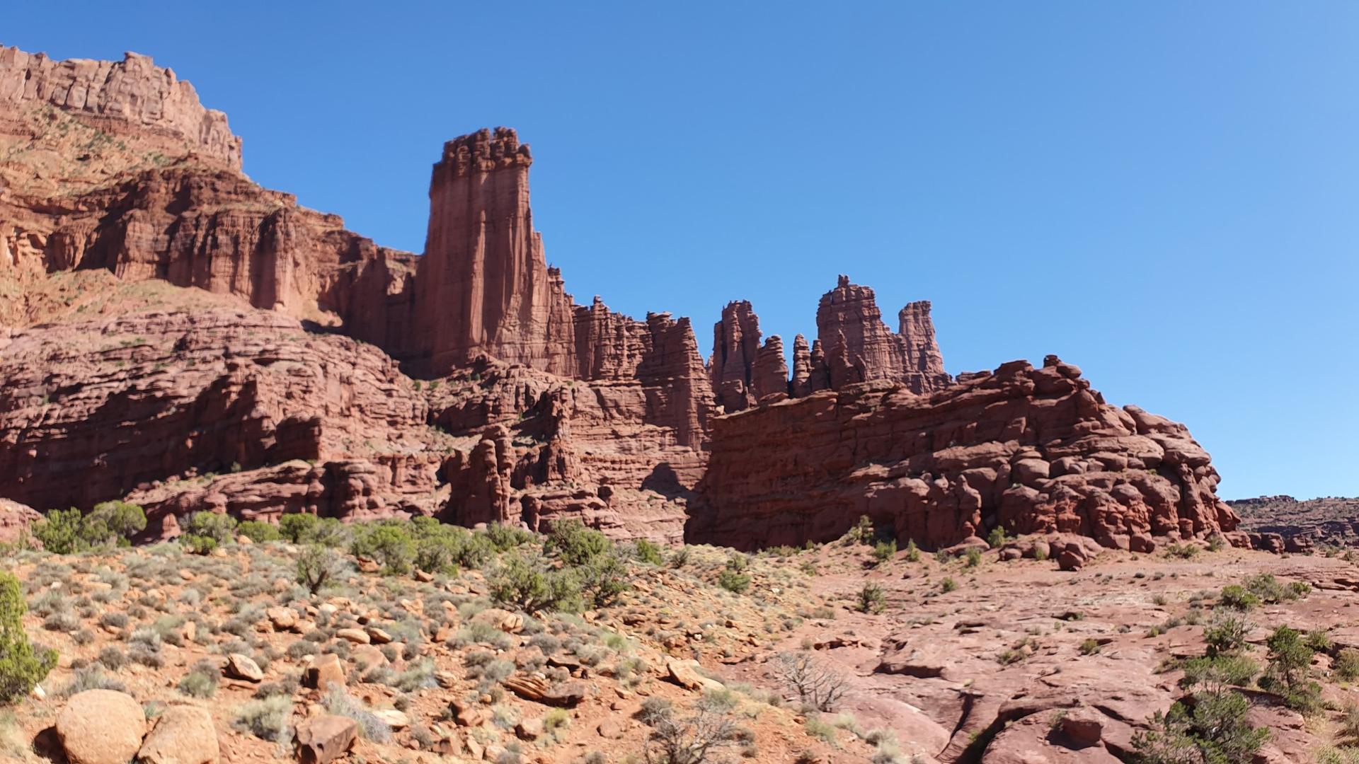 Wide view of multiple red rock formations and desert shrubs under a clear blue sky.