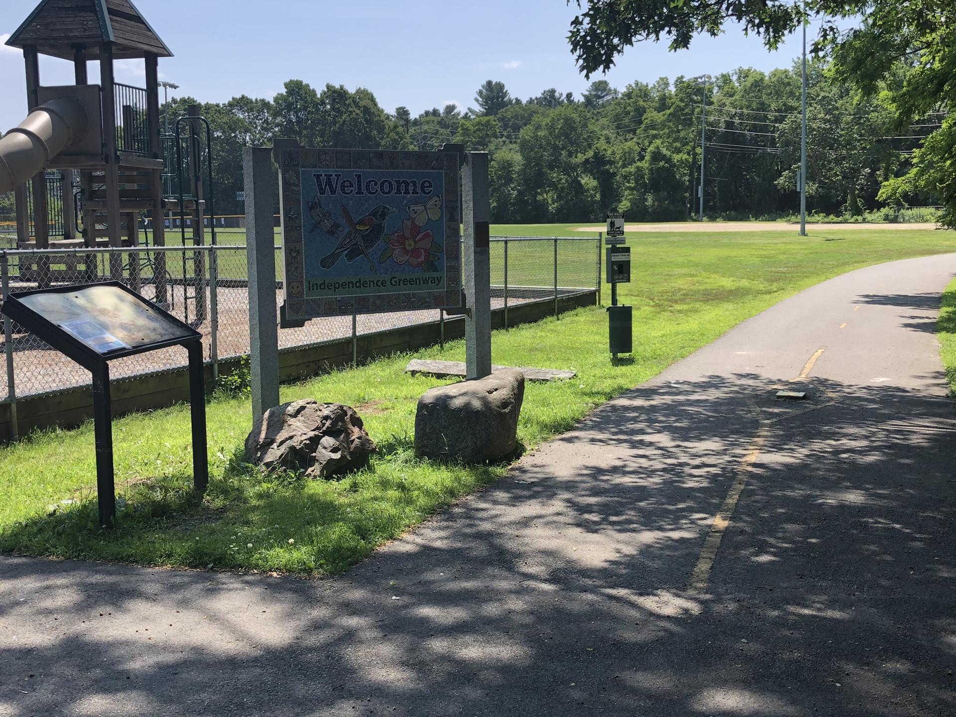 Paved path to a playground with a welcome sign and green field.