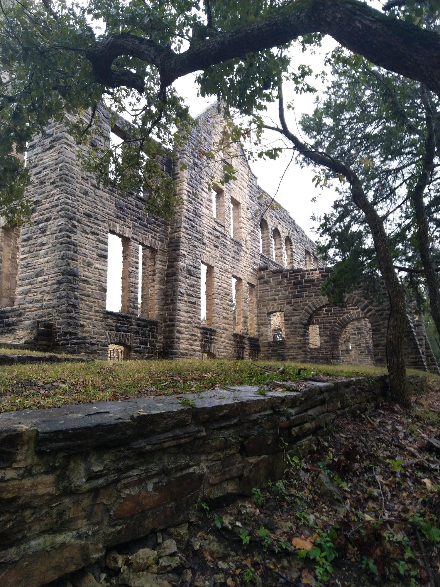 Stone ruins of a large building on a grassy hill, framed by trees.