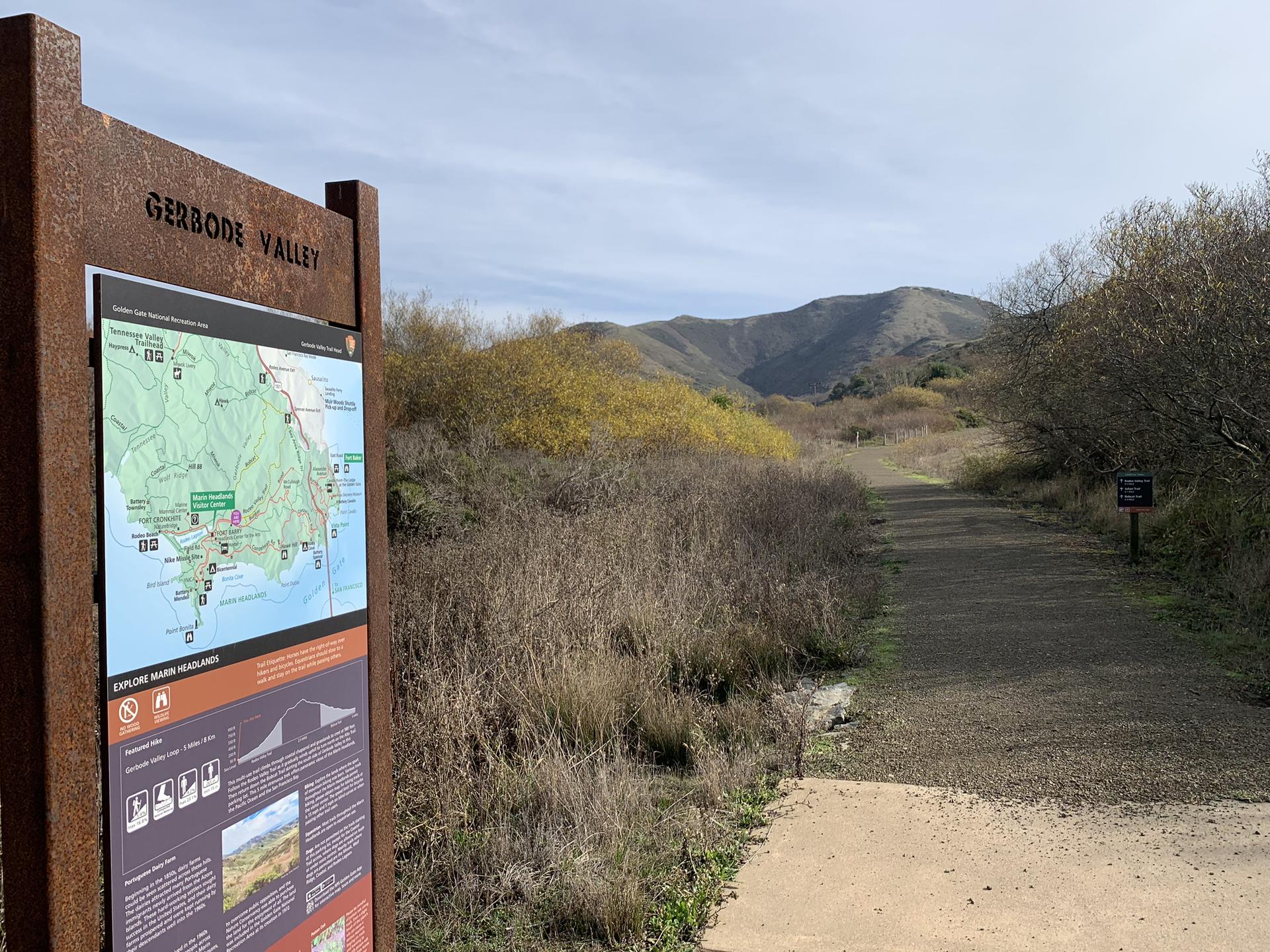 A trail sign next to a dirt path leading into a valley with hills.