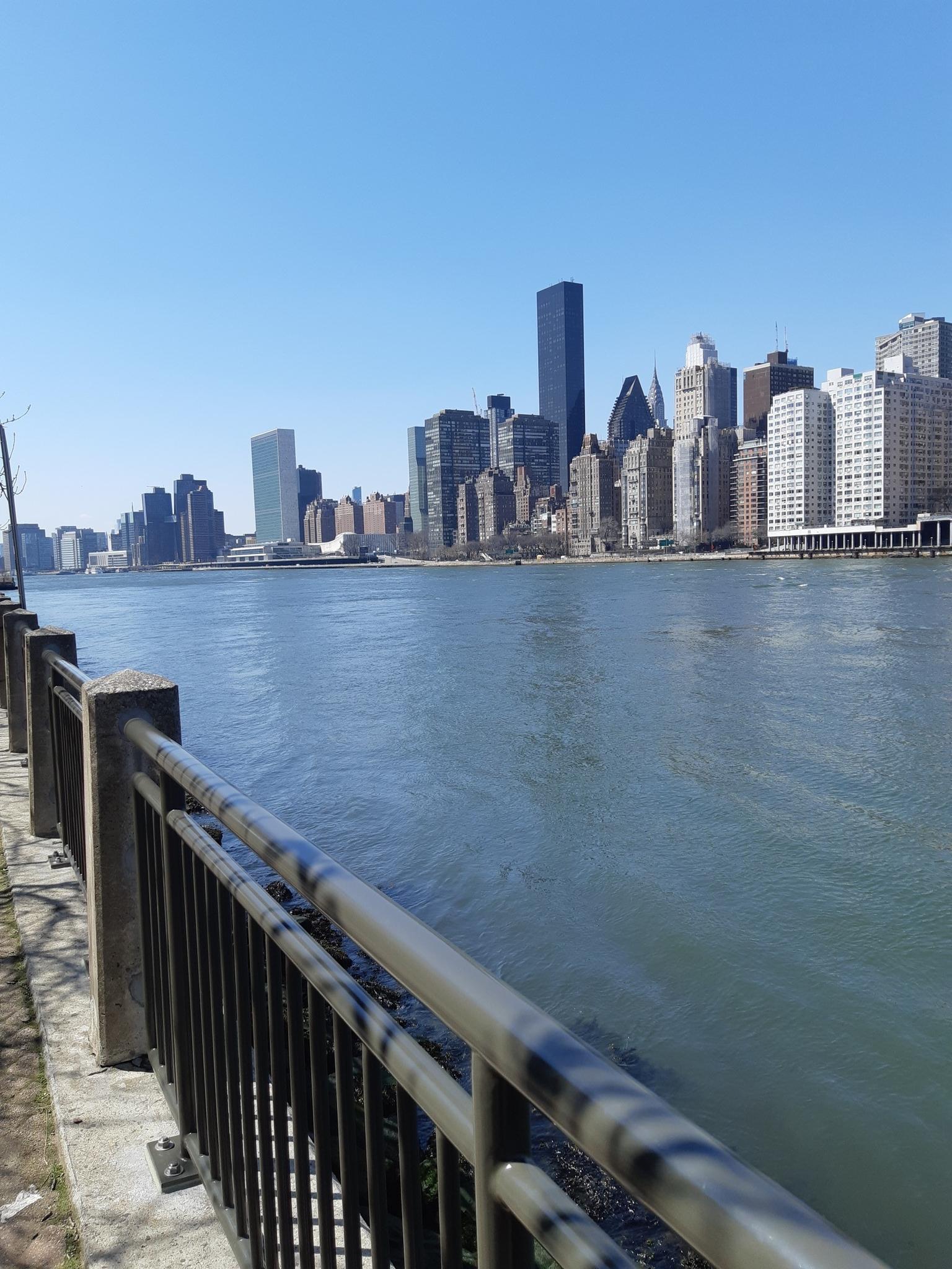 City skyline across a river, viewed from a fenced waterfront path on a sunny day.