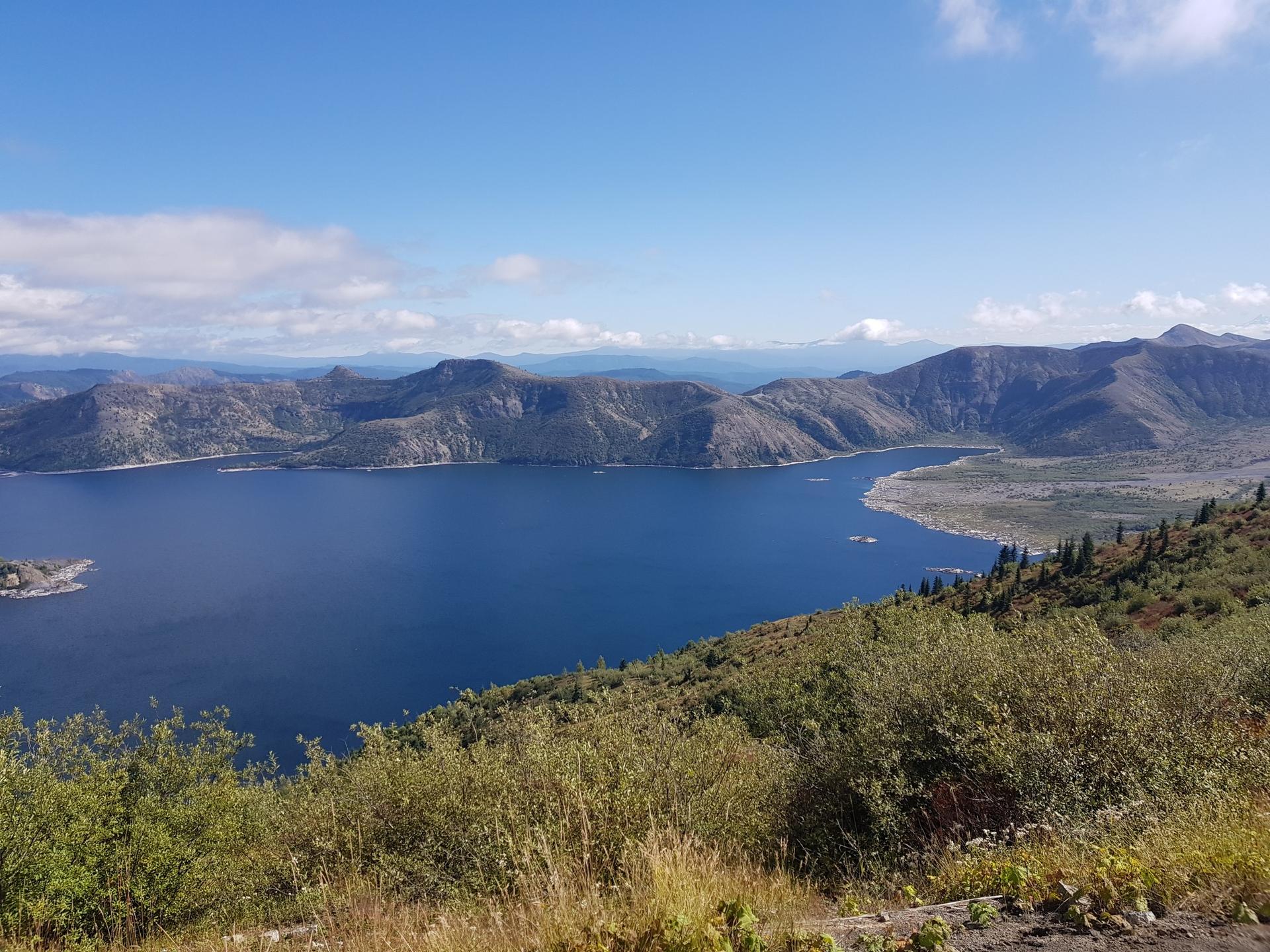 Sunny panoramic view of a vast blue lake surrounded by green mountains.