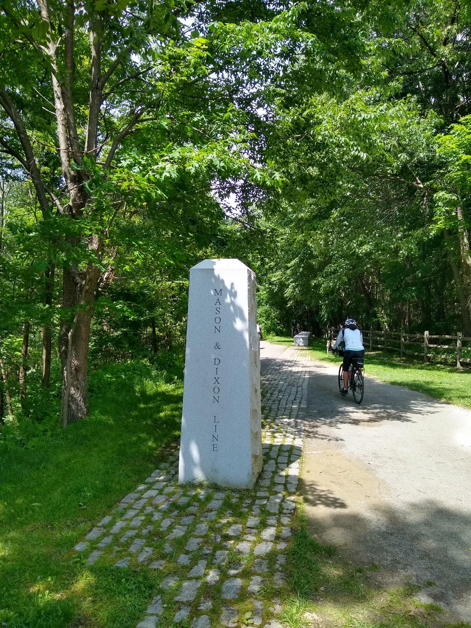 A cyclist rides past a tall stone monument on a sunny, tree-lined path.