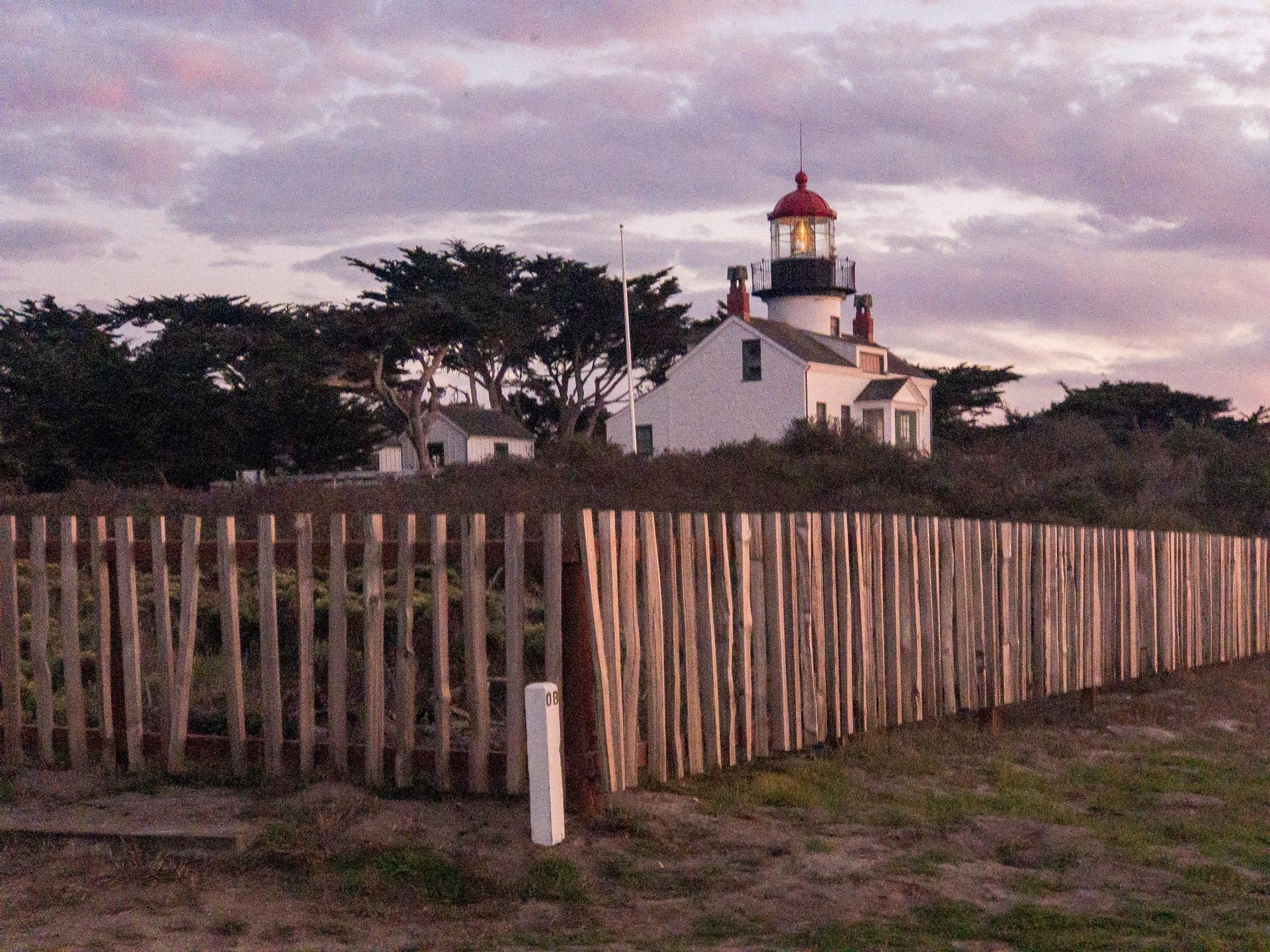 White lighthouse with red top behind a wooden fence at dusk.