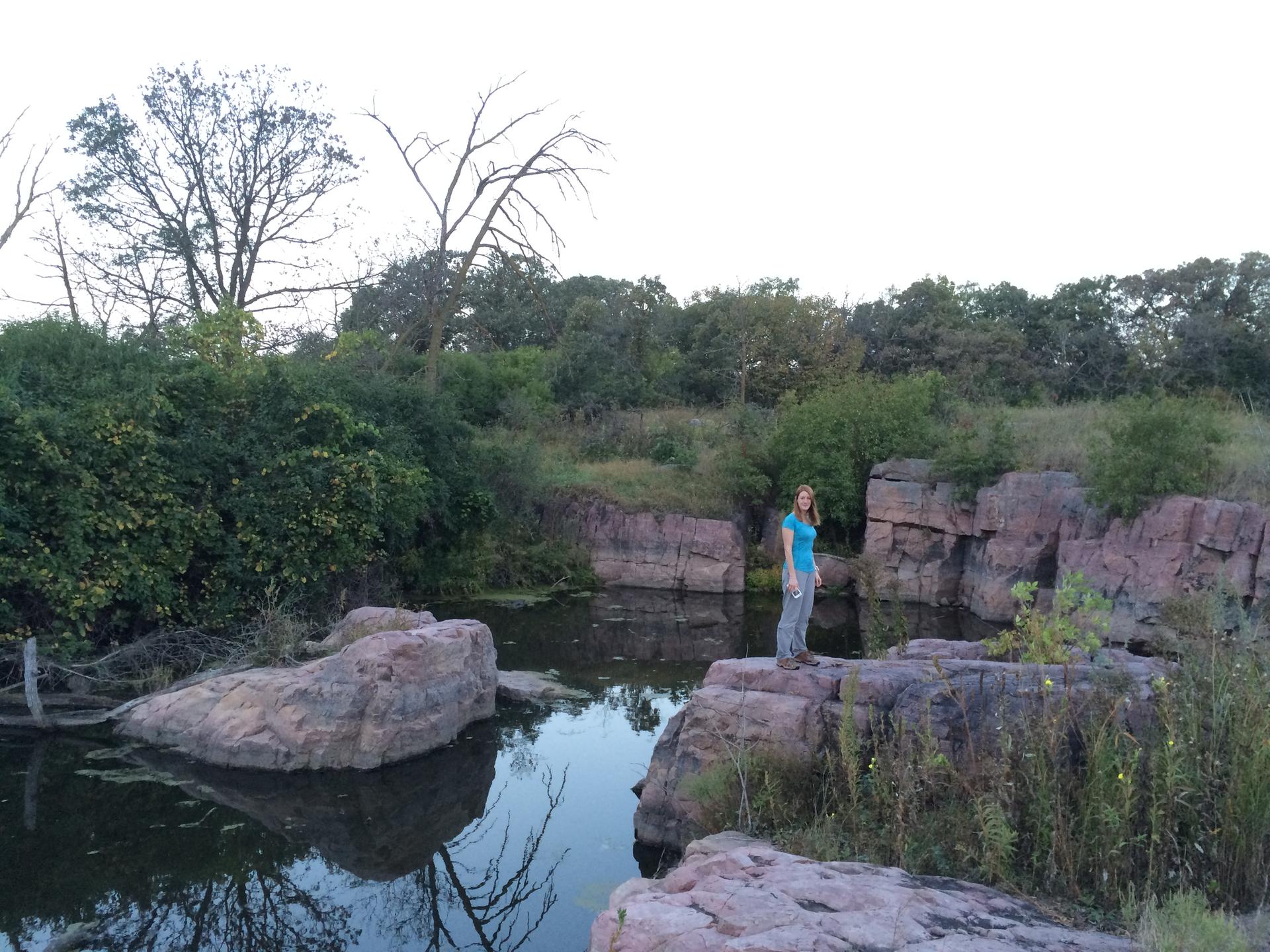 Woman on a rock by calm water, surrounded by rocky cliffs and trees.