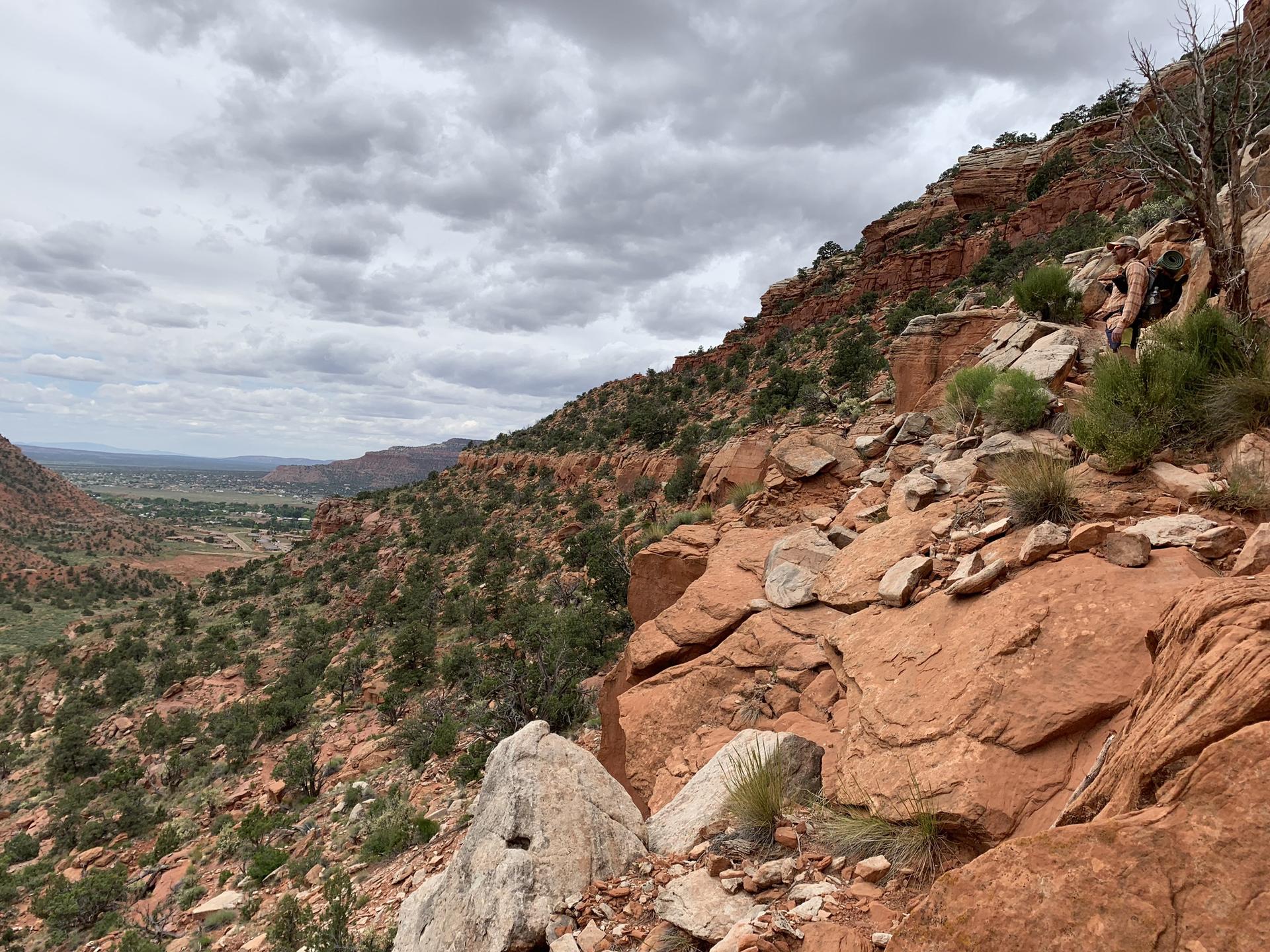 Person hikes up a steep, rocky red hillside overlooking a green valley.