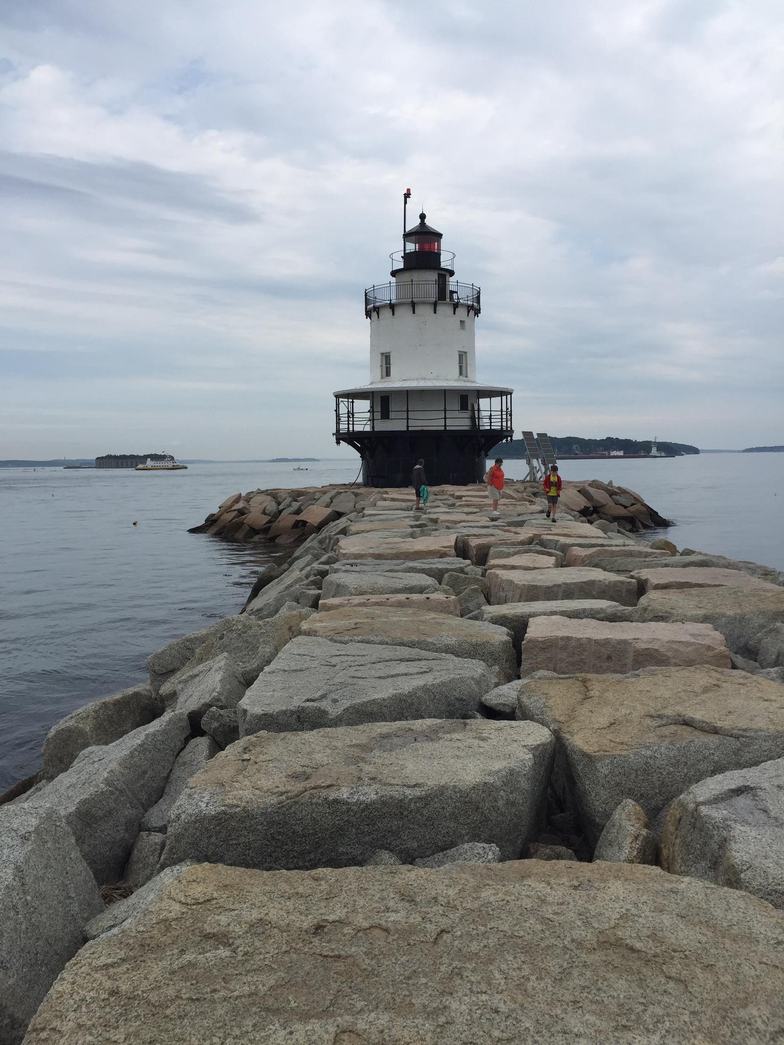 People walk on a rocky jetty towards a white lighthouse under an overcast sky.