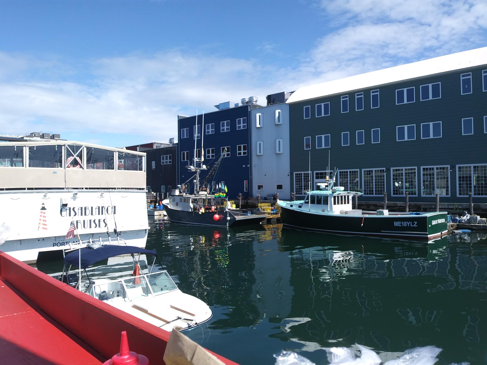 Multiple boats docked in calm water next to large waterfront buildings on a sunny day.