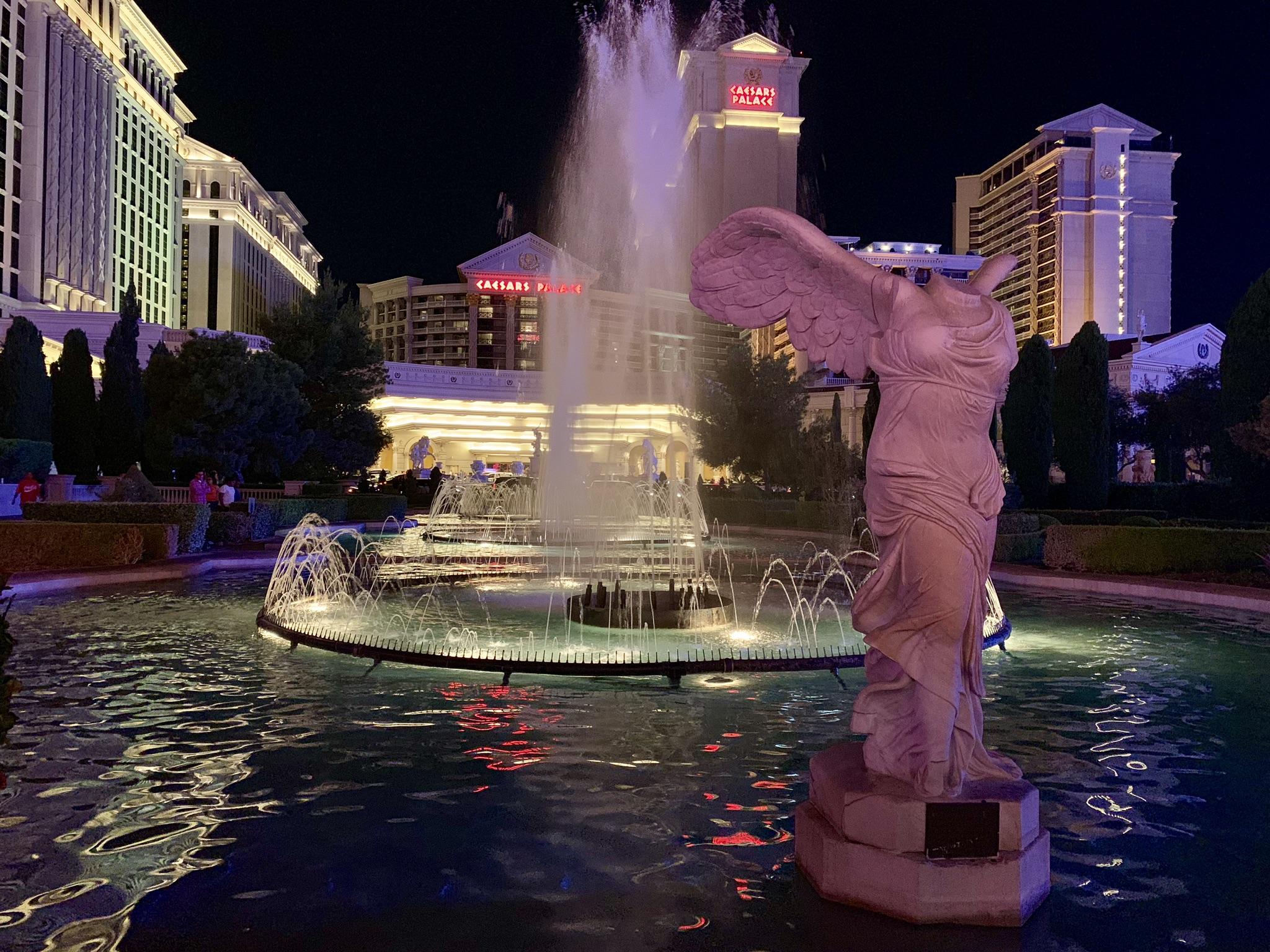 Statue and illuminated fountain at night with distant buildings glowing.