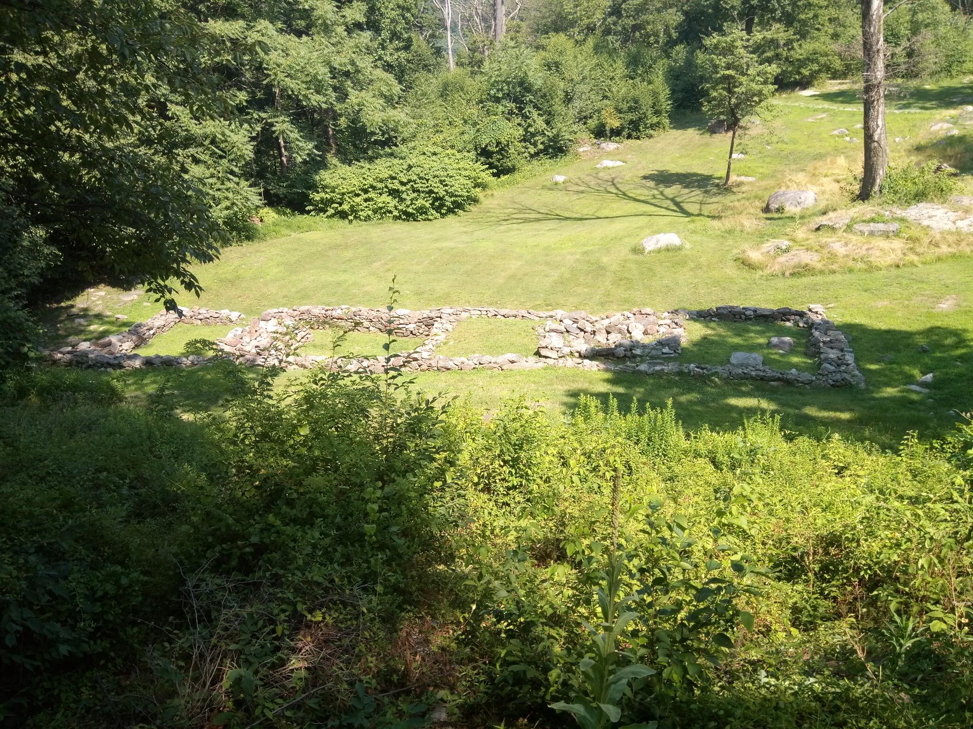 Stone foundation ruins in a sunny, grassy clearing surrounded by trees.