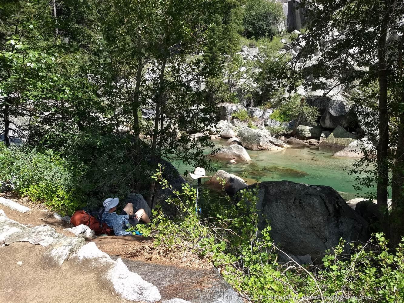 Person resting by a clear, shallow river with large rocks and green trees. Sunny.