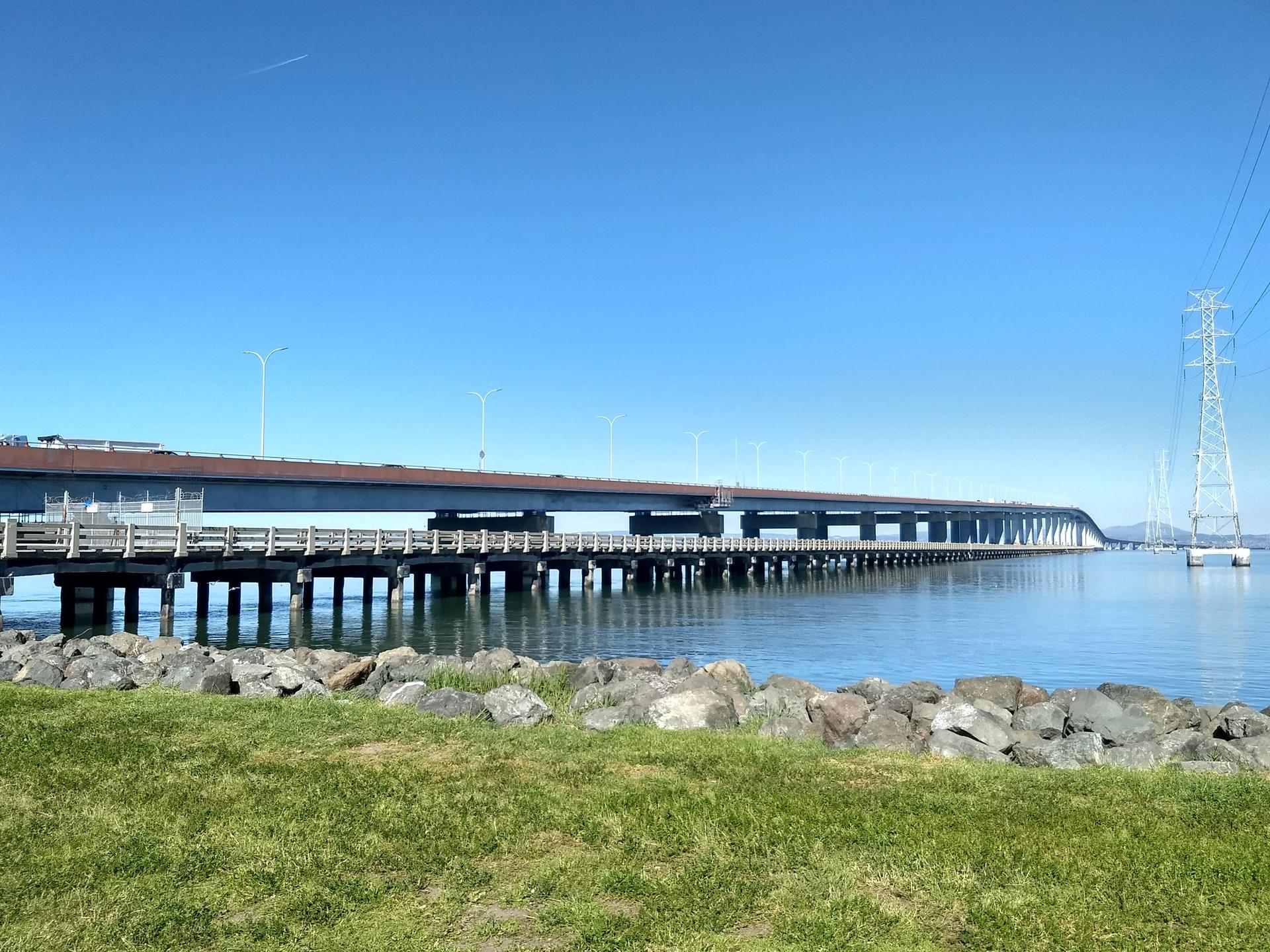 Long bridge over calm water with power lines and green grass in the foreground.