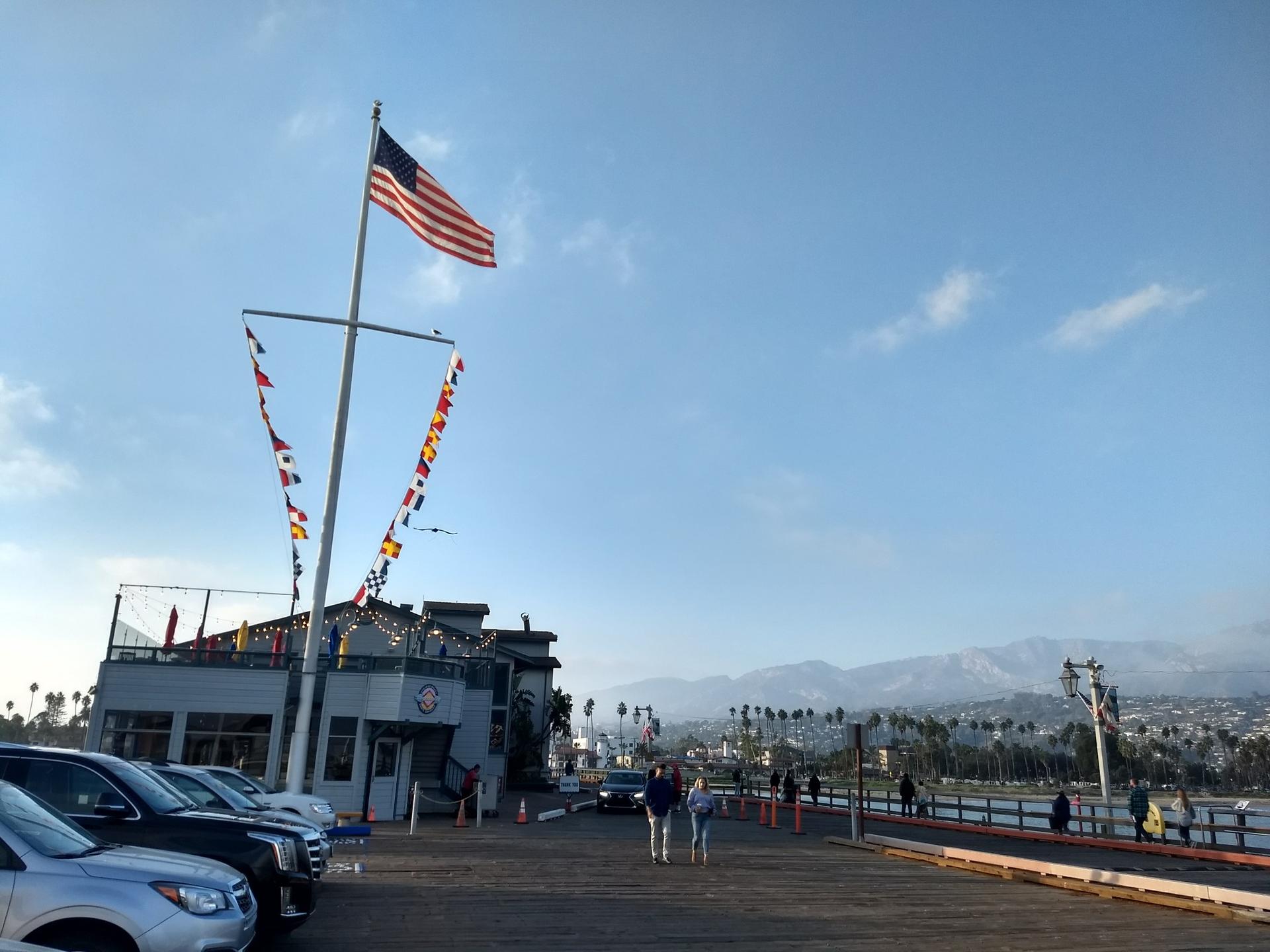 American flag on pier building with parked cars and distant mountains.