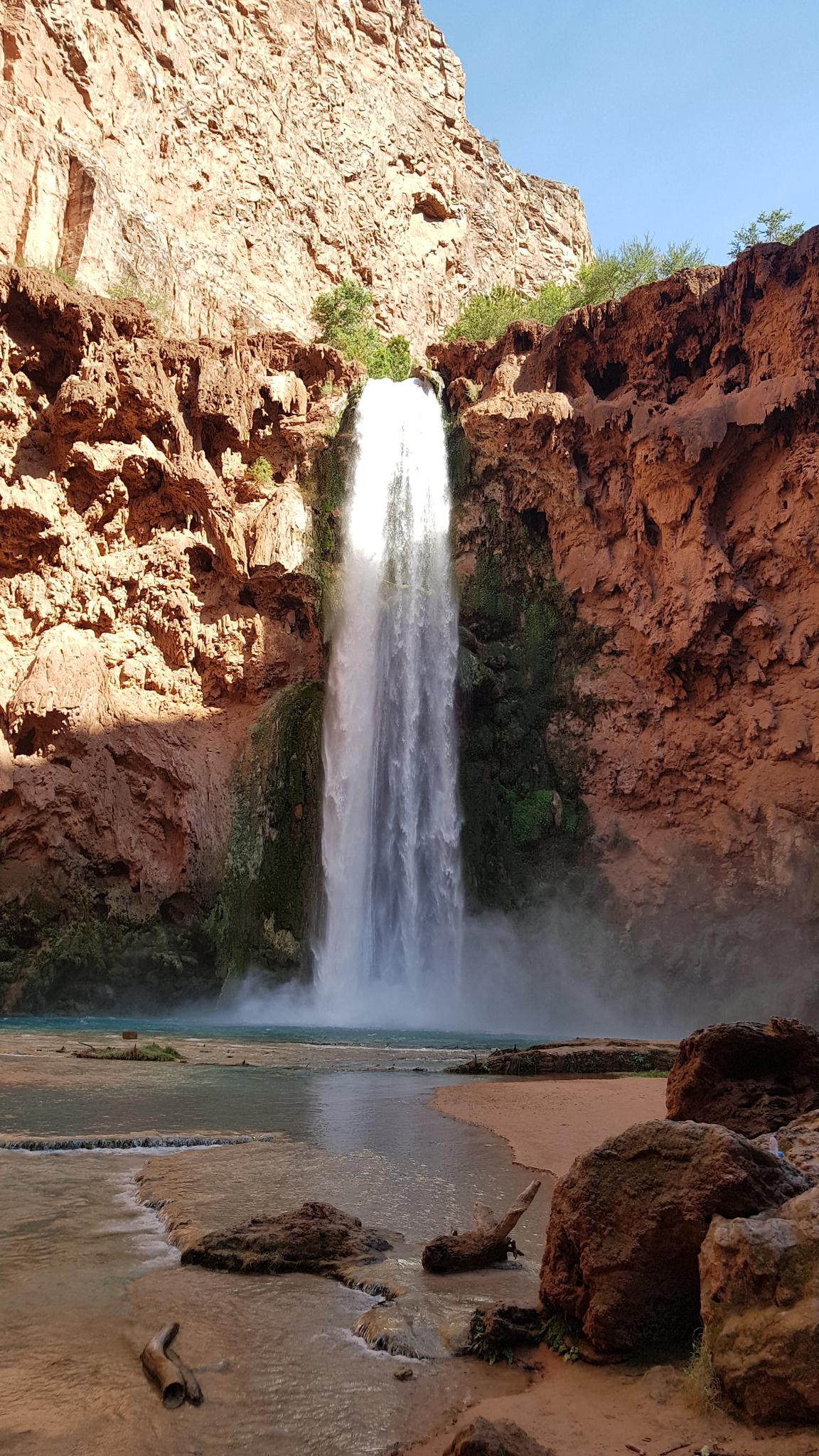 A tall waterfall plunges into a turquoise pool at the base of a red canyon.