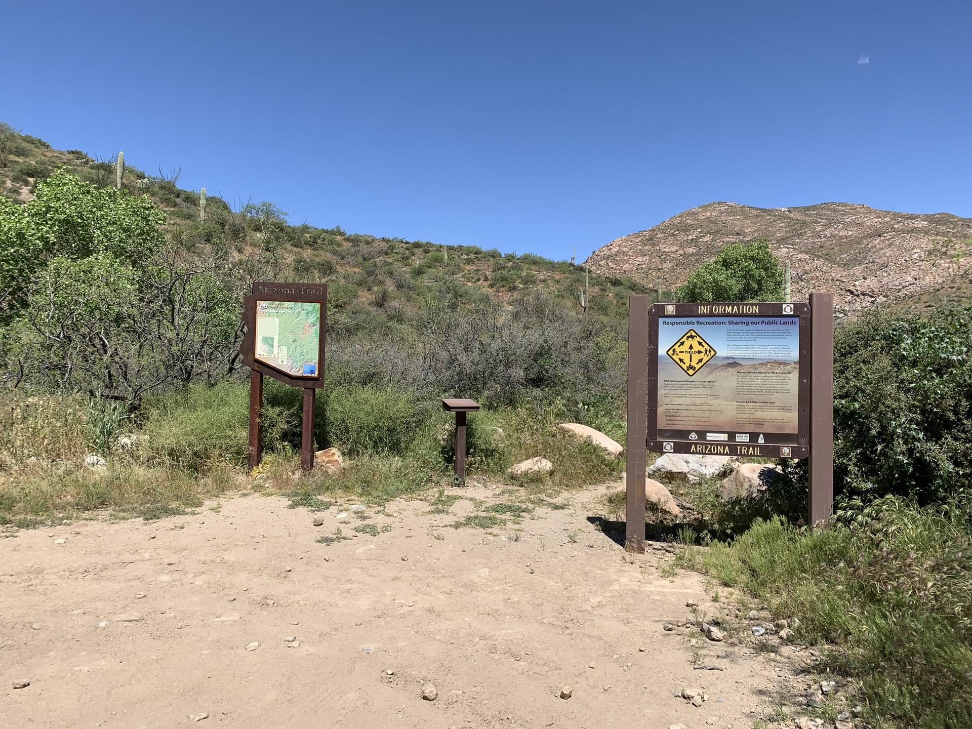 Two information signs on a dirt path surrounded by dry brush and hills.