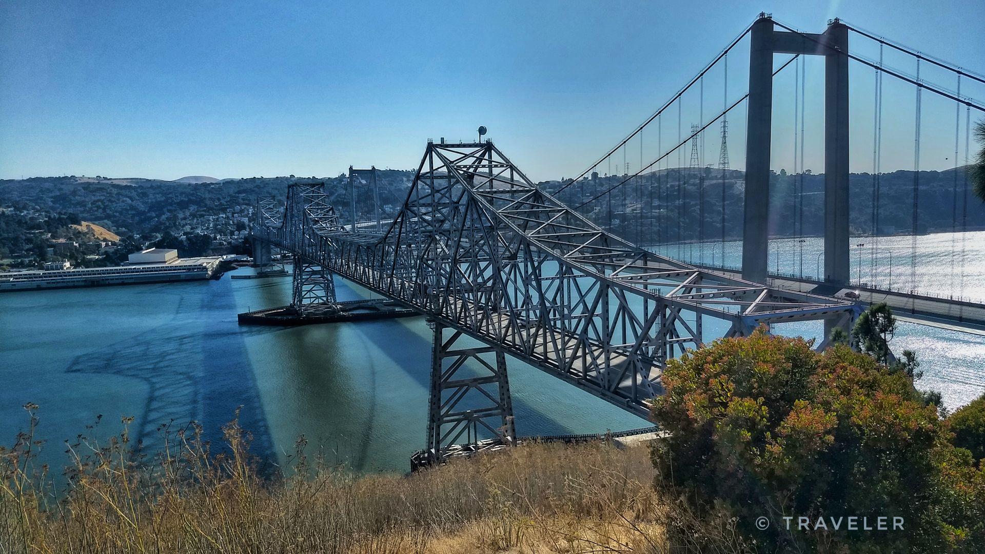 A massive metal bridge crosses wide water on a sunny day.