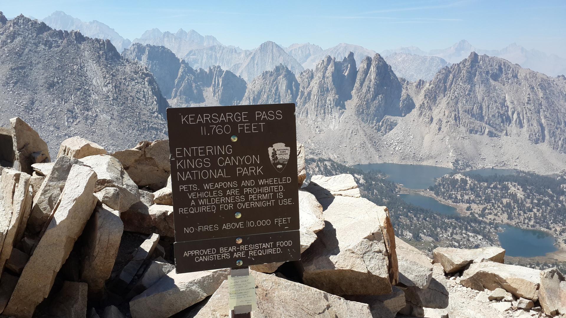 A wooden sign on a rocky mountain pass overlooks distant mountains and lakes.