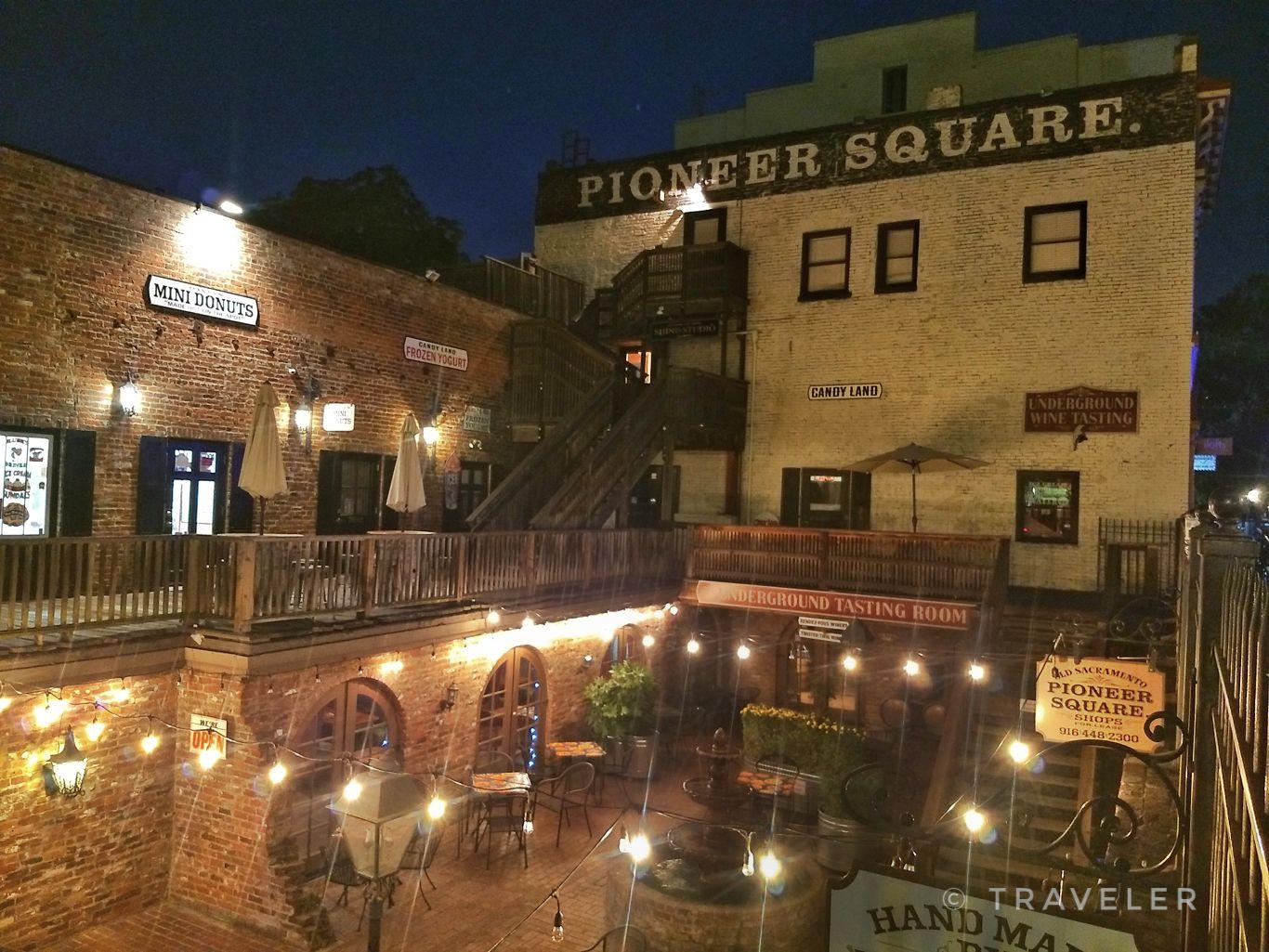 Old brick buildings with string lights illuminating an outdoor seating area at night.