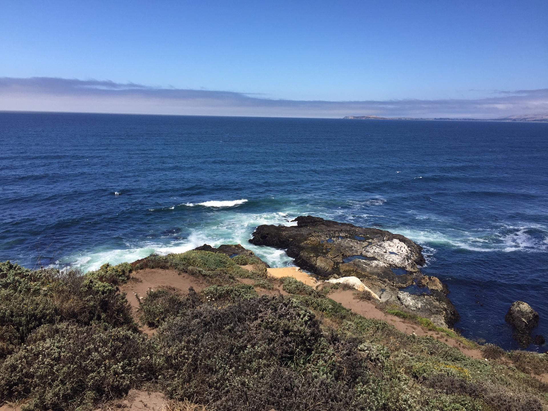 Deep blue ocean waves crash against dark rocky outcrops and green coastal cliffs.