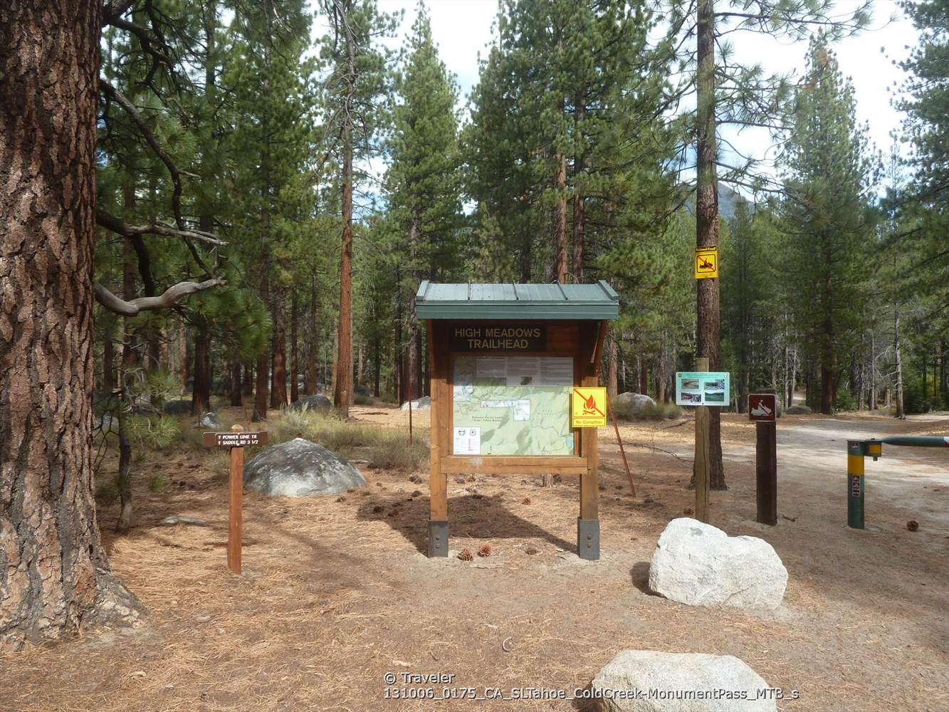 A trailhead sign with maps stands among tall pine trees in dappled sunlight.