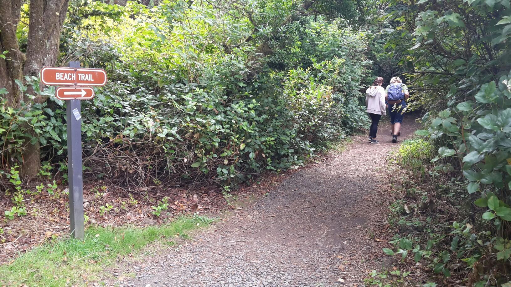 Two people walk on a gravel path through dense green foliage, past a trail sign.