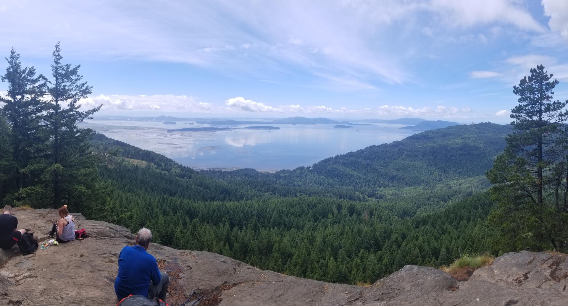 Panoramic view of a vast lake and mountains under a cloudy sky, with people resting.