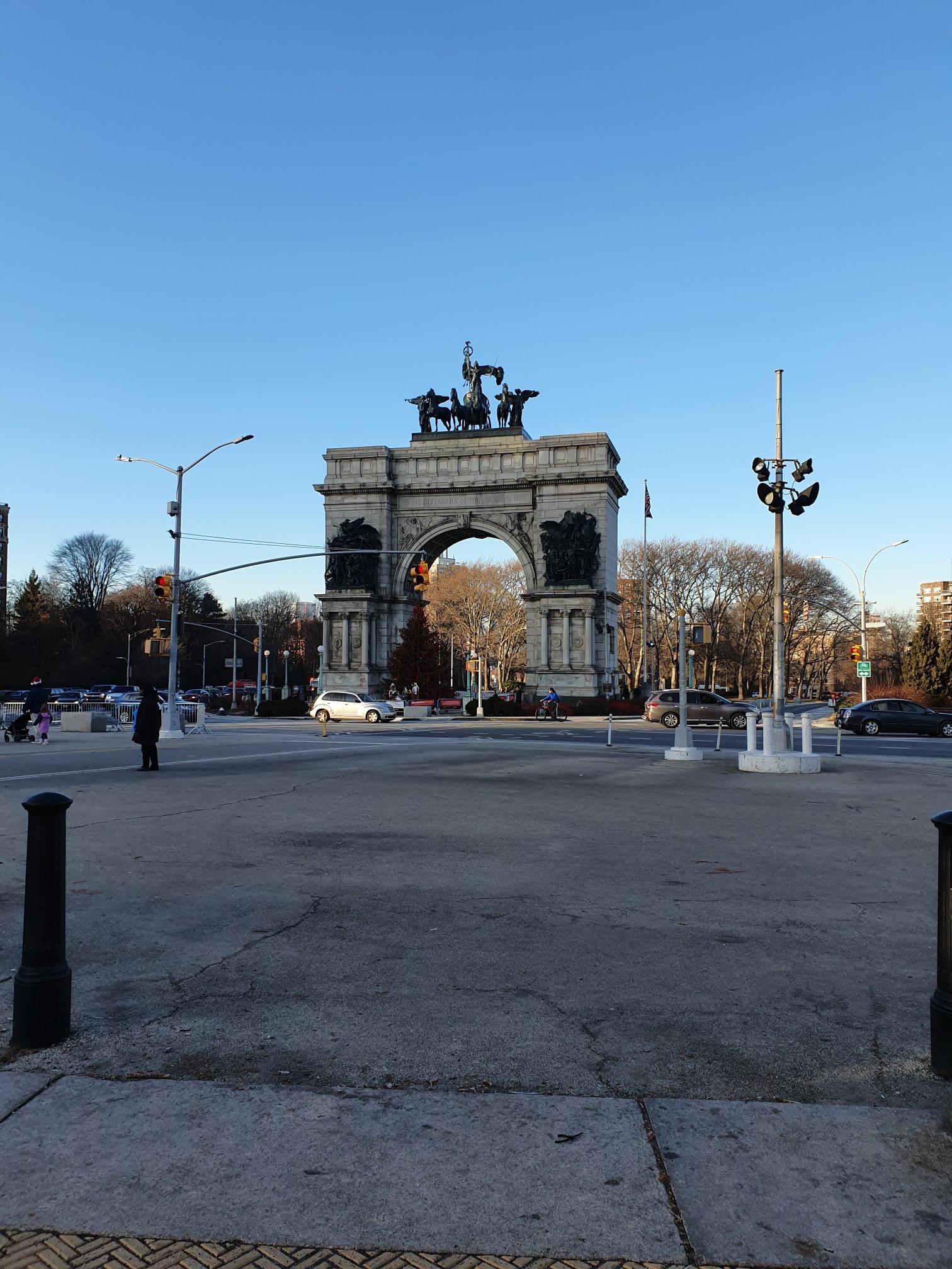 Large stone archway with statues under a clear blue sky, cars nearby.