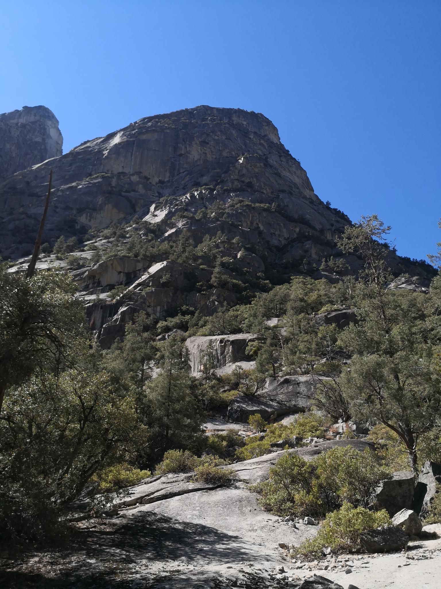 Steep, rocky mountain rising under a clear blue sky, with sparse trees.