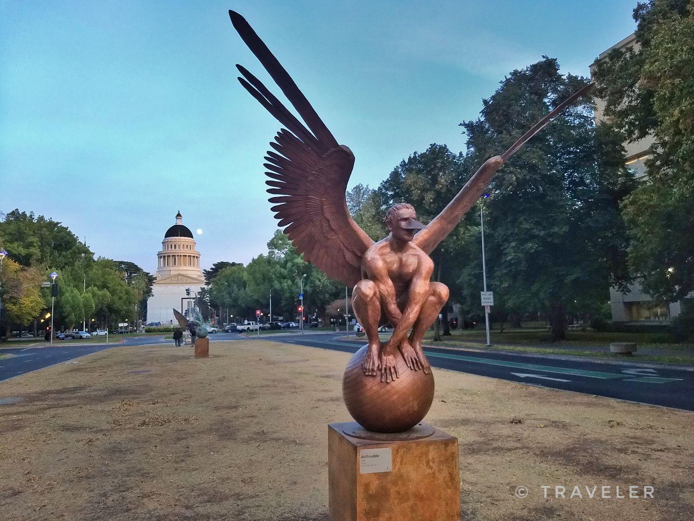 Large winged statue overlooking a domed building and park at dusk.