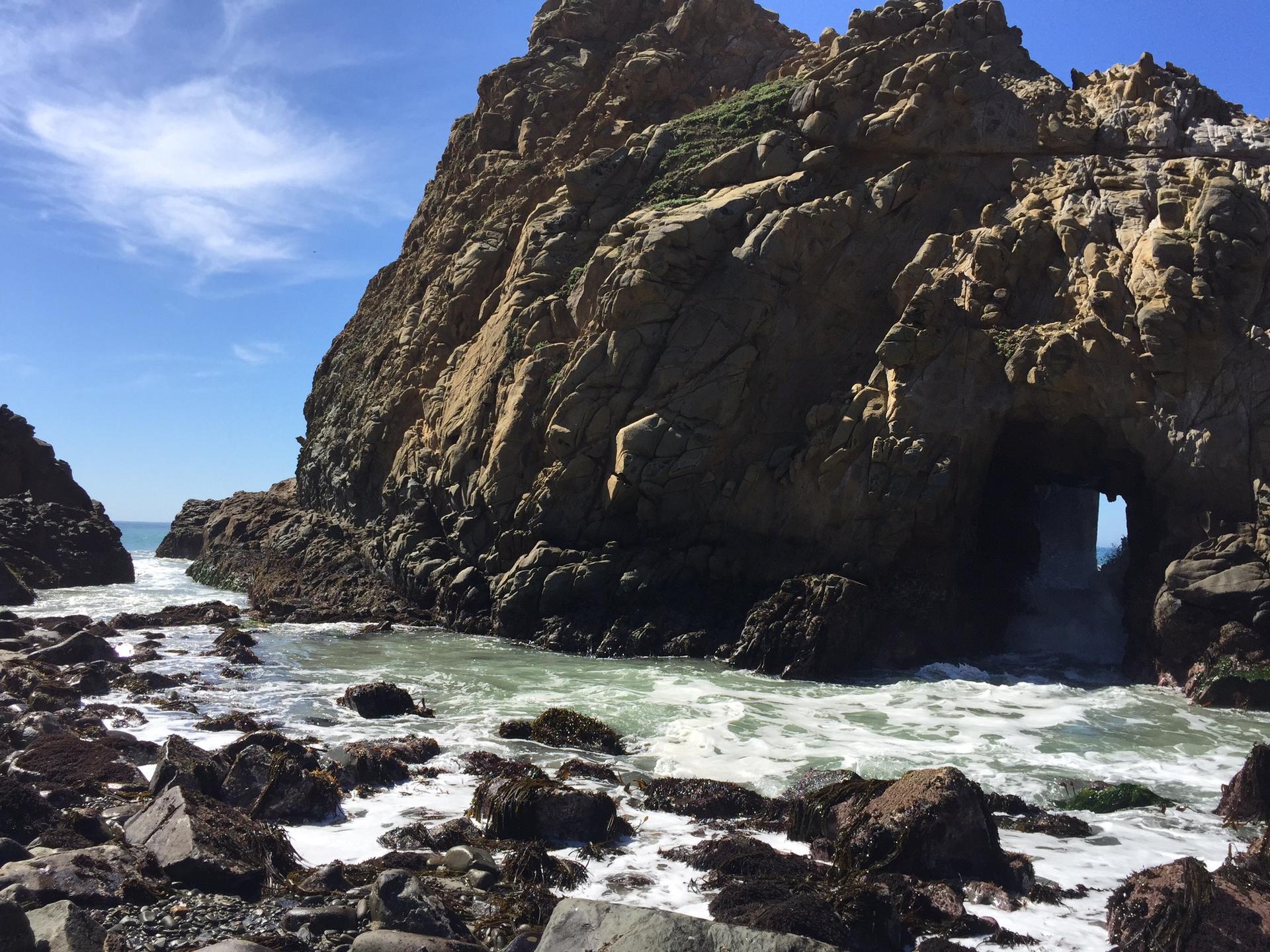 Sunny view of a large rocky sea stack with an archway and foamy water.