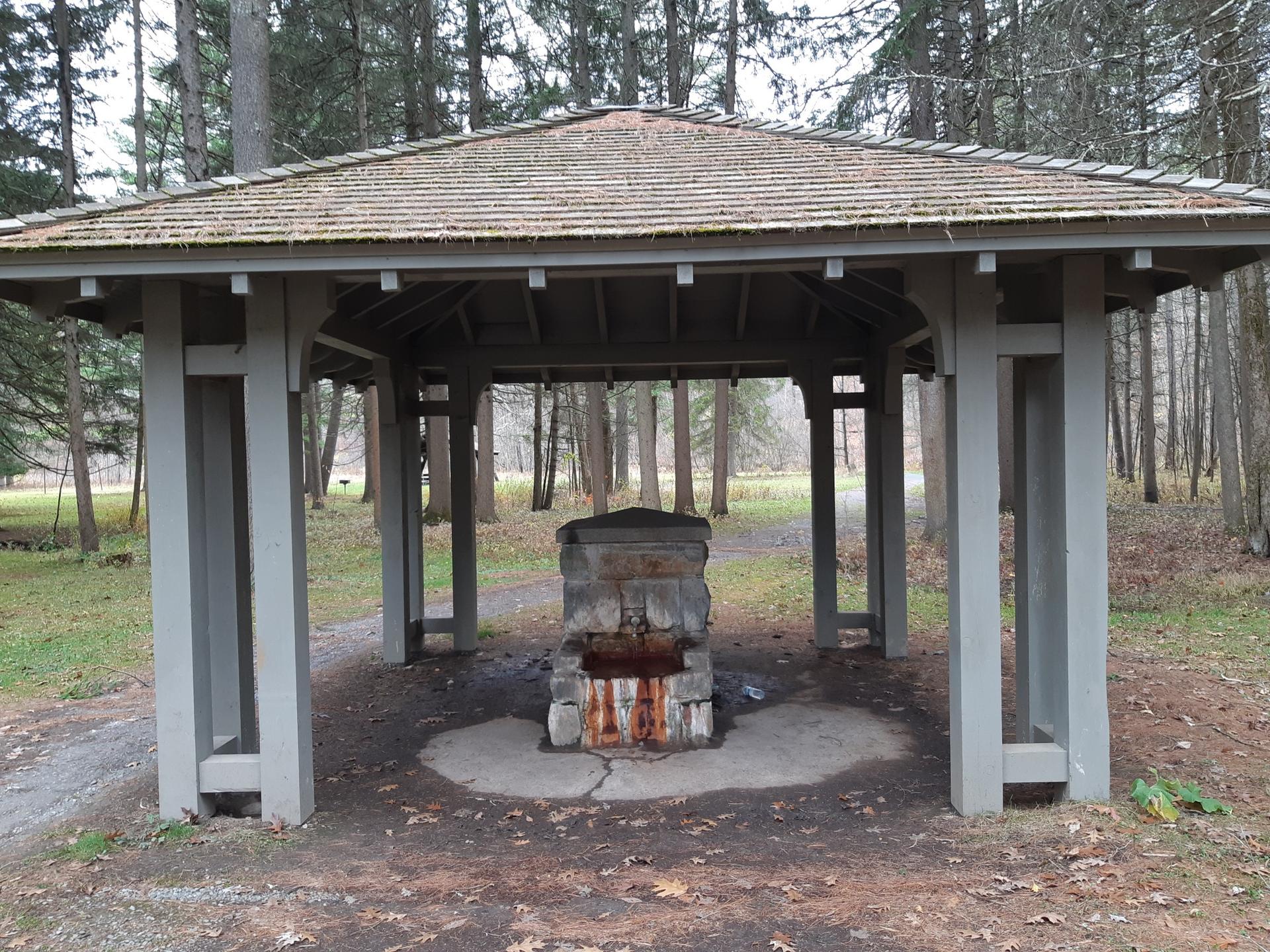 Wooden gazebo sheltering an old stone structure in a wooded park.