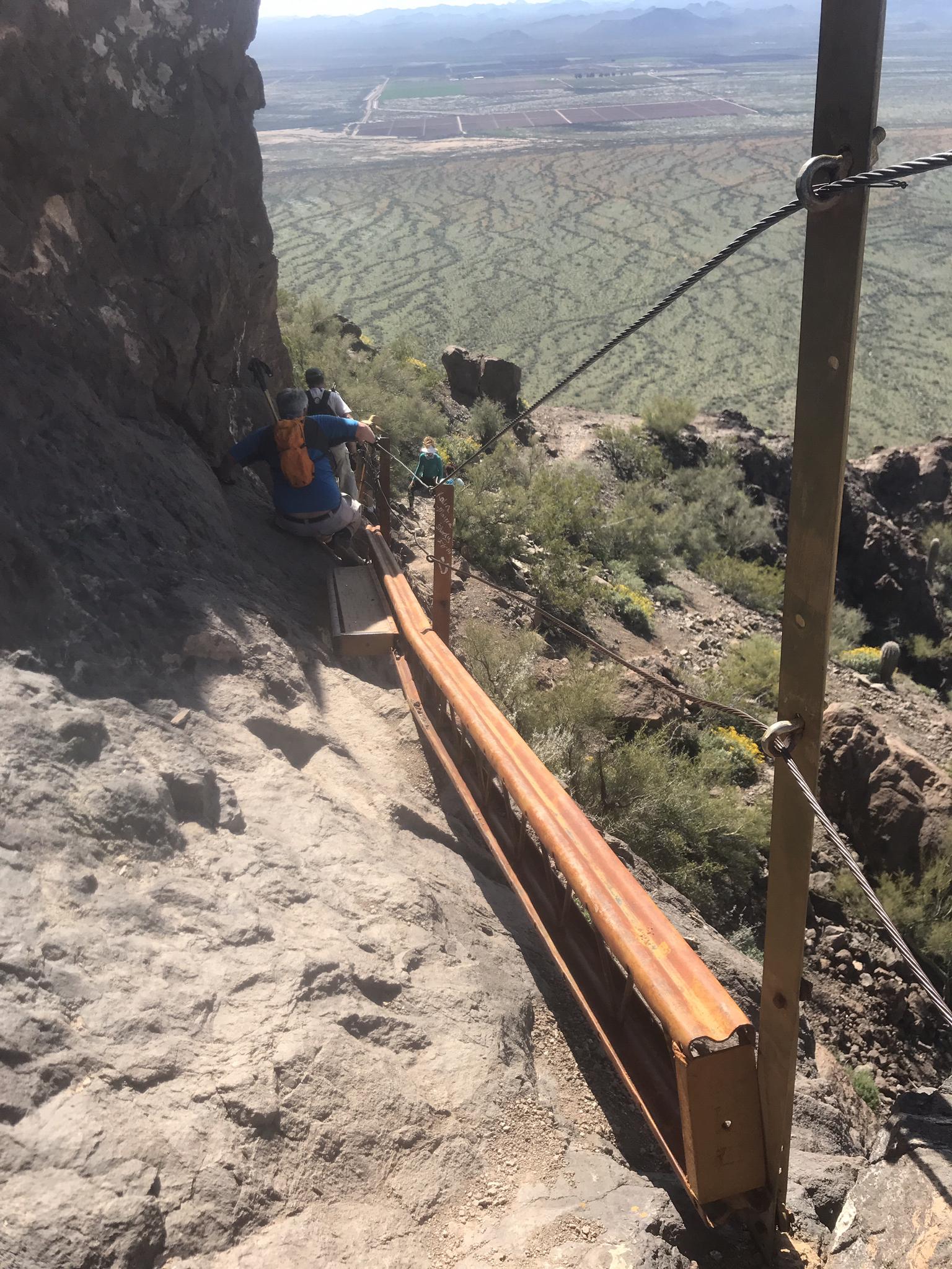 Hiker on a narrow cliffside trail with safety cables overlooking a vast desert.