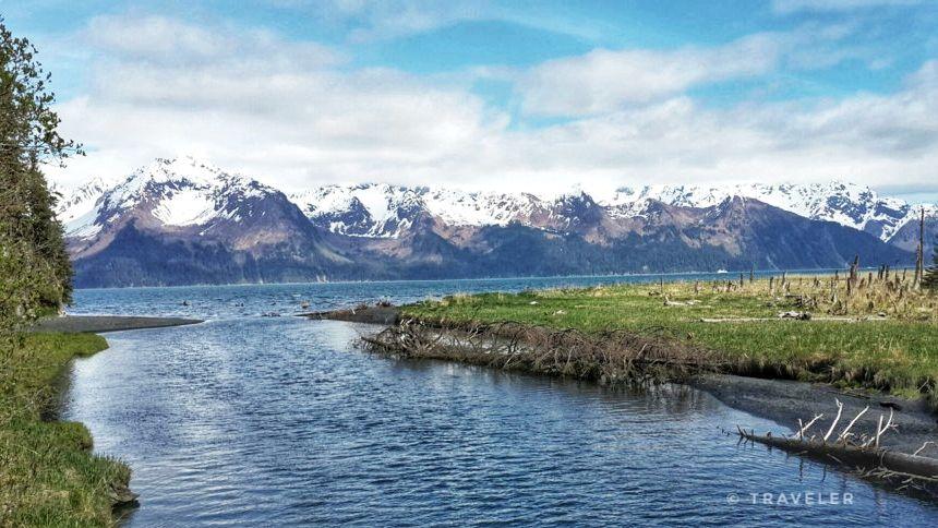 River flowing into a large body of water with snow-capped mountains and cloudy sky.