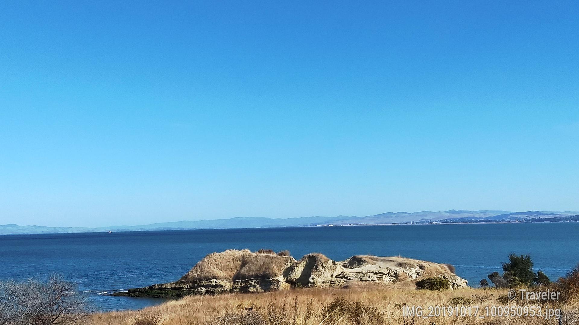 Bright sunny day over blue water with a small rocky island and distant land.