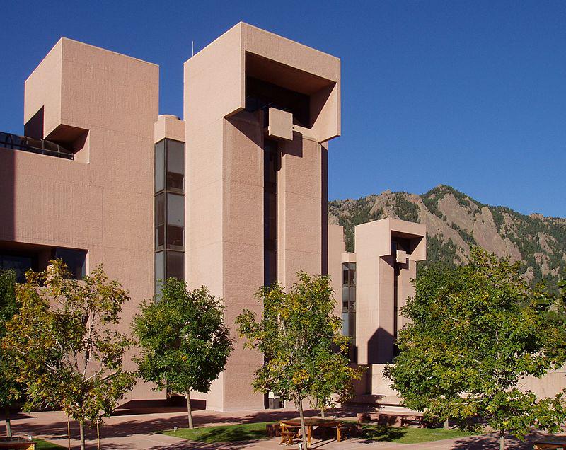 Modern building with geometric shapes, trees, and mountains under clear sky.