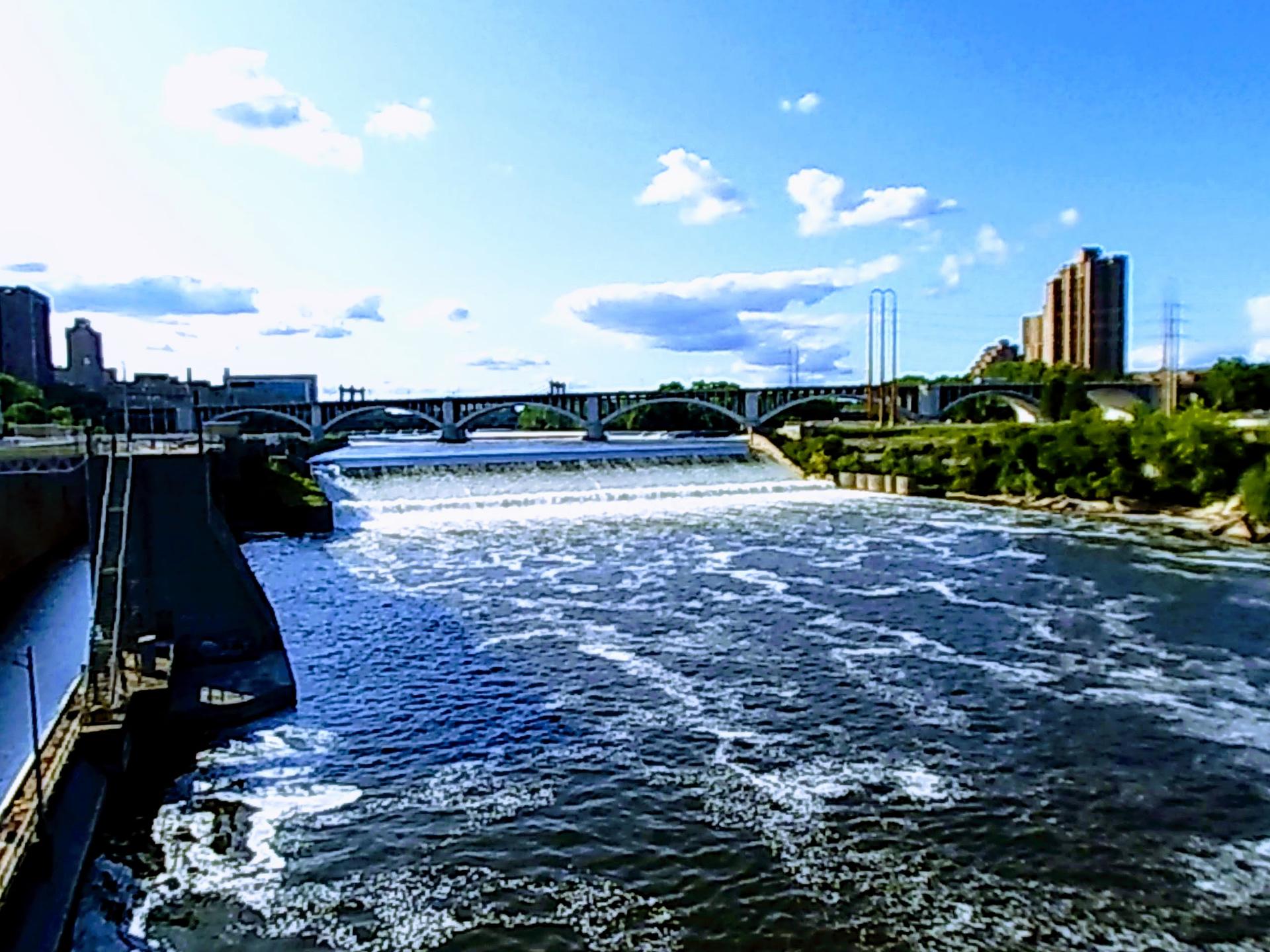 Wide river with a dam and foamy water, city buildings, and bridges under a bright sky.