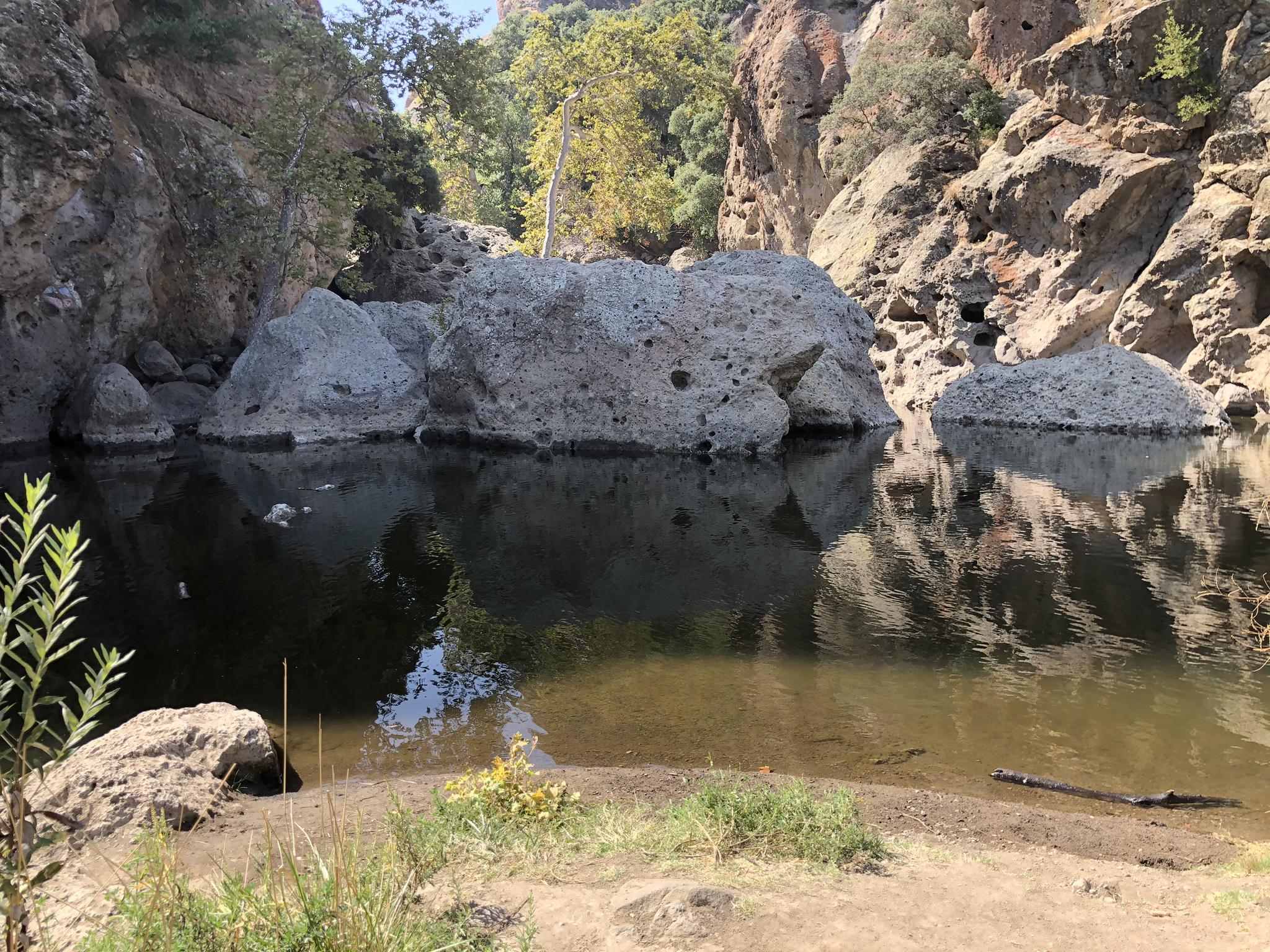 Calm water reflecting large rocks and green trees under bright sky.