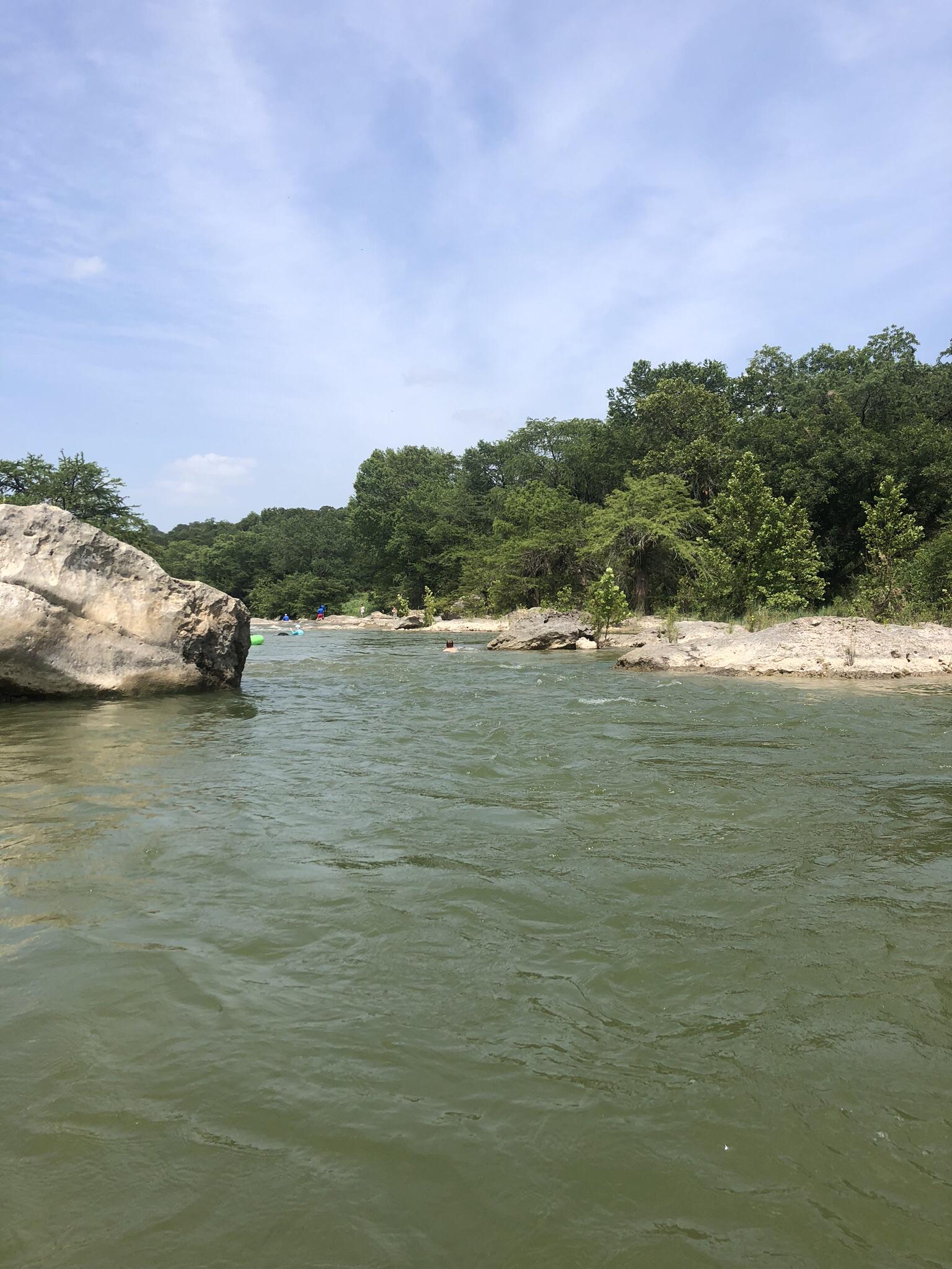 Wide green river flowing past a large rock and lush trees on a bright day.