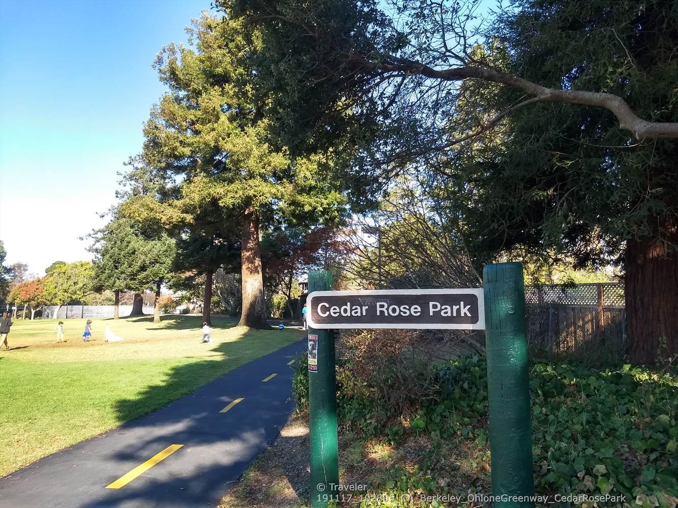 A park sign next to a paved path with green grass and tall trees.