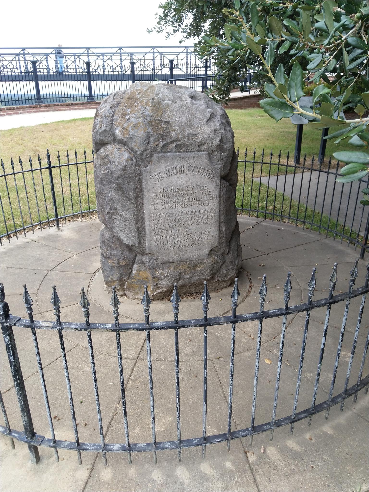 Large stone monument with an inscription, surrounded by a black metal fence.
