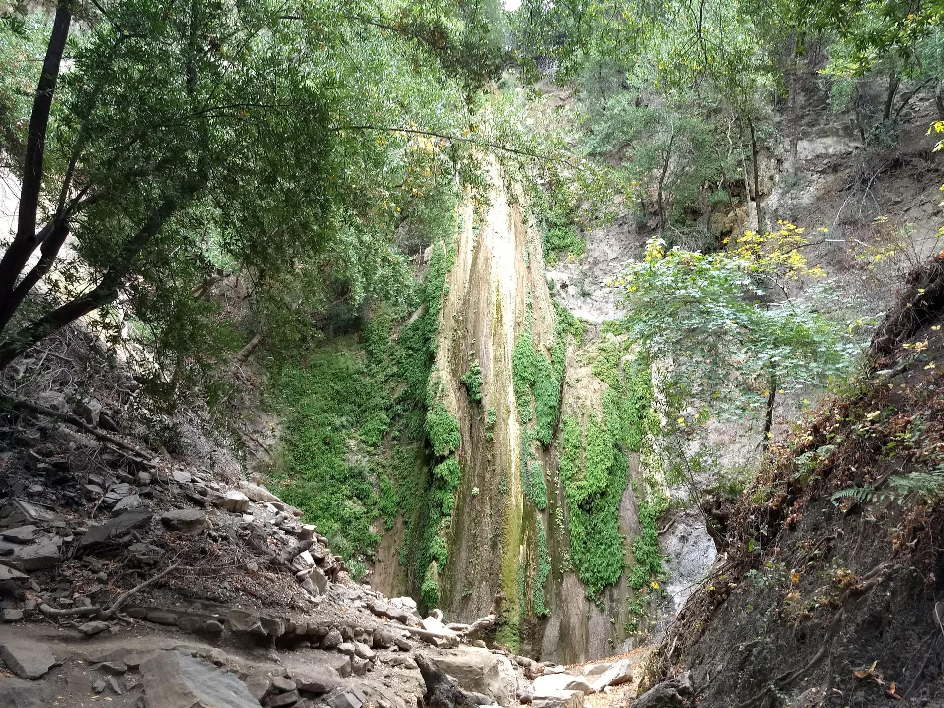 A tall, moss-covered waterfall cascades down a rocky cliff in a green forest.
