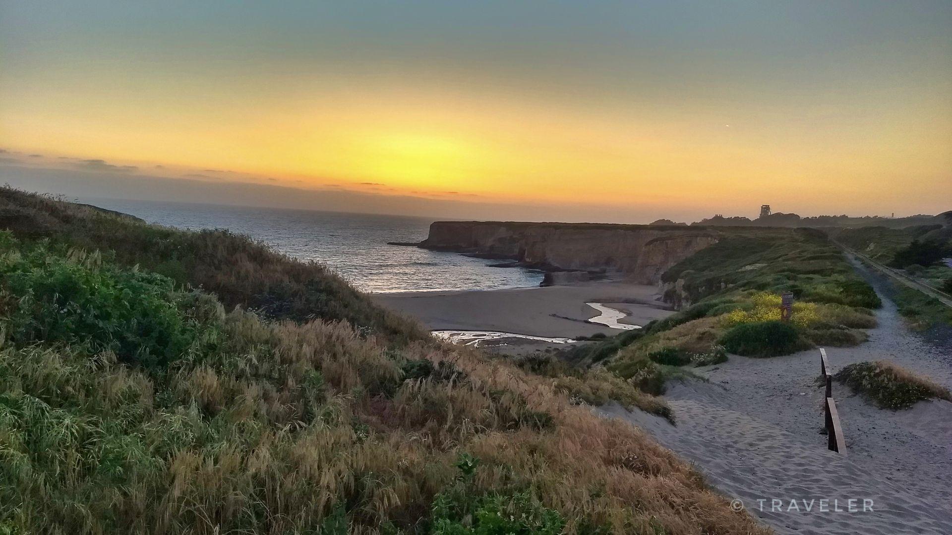 Coastal sunset over a sandy beach with cliffs and a surfboard.