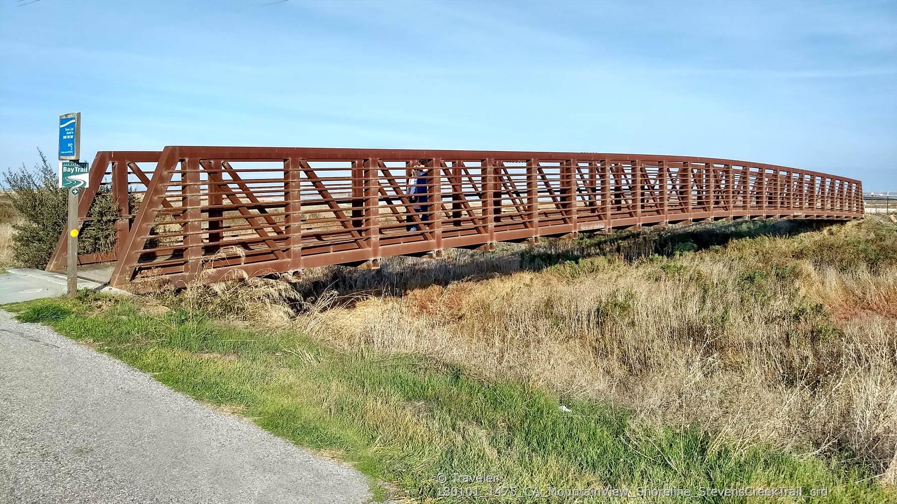A person walks across a long, rusty metal bridge over dry grass under a clear sky.