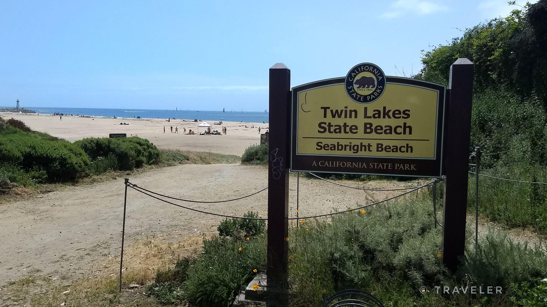 A wooden sign on a sandy path leads to a sunny beach with distant people.